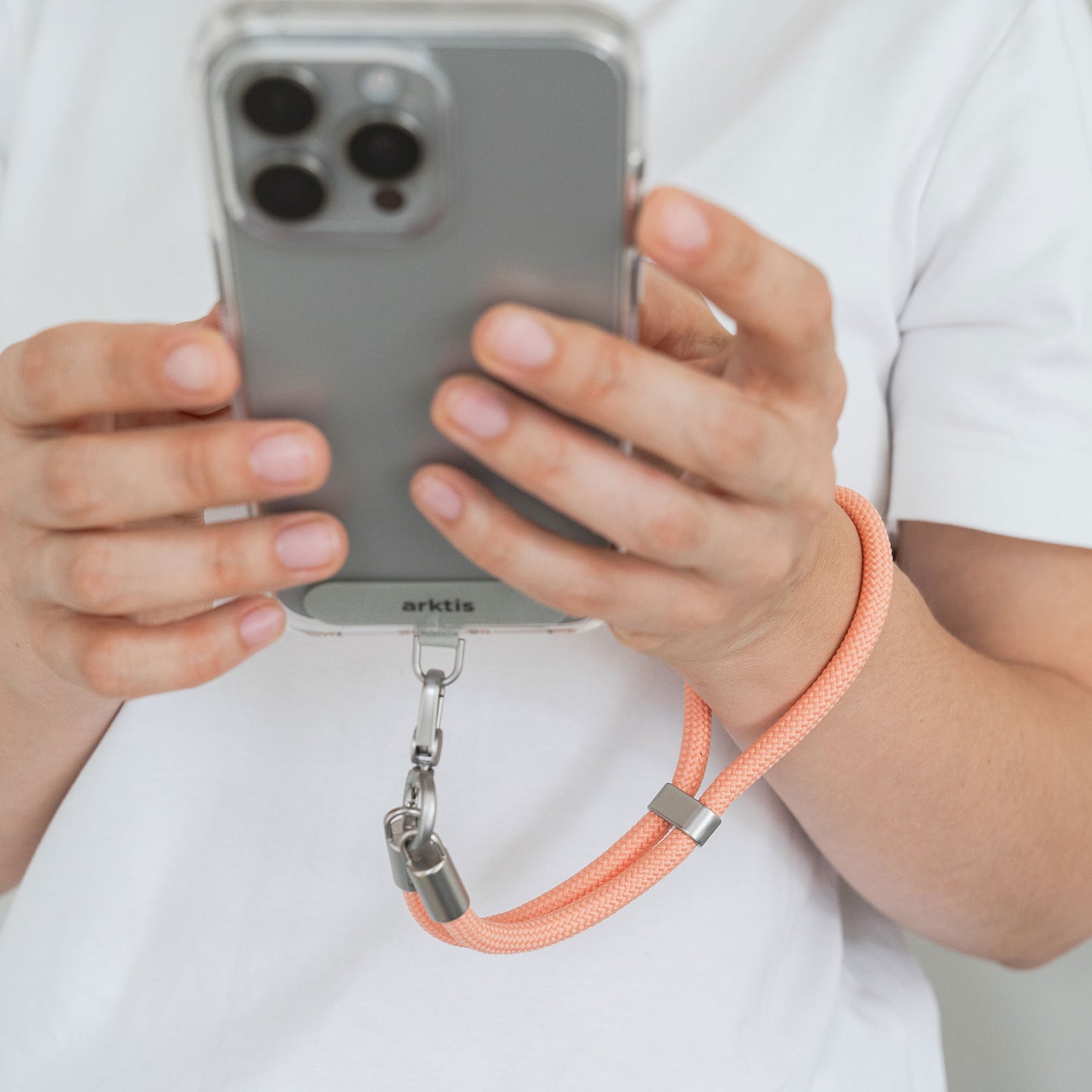 Person, die ein Telefon mit der arktis Lanyard Handykette fürs Handgelenk von Arktis in Pfirsich in der Hand hält, eine stilvolle Mischung aus Smartphone-Zubehör und Mode-Accessoire, gepaart mit einem weißen Hemd.