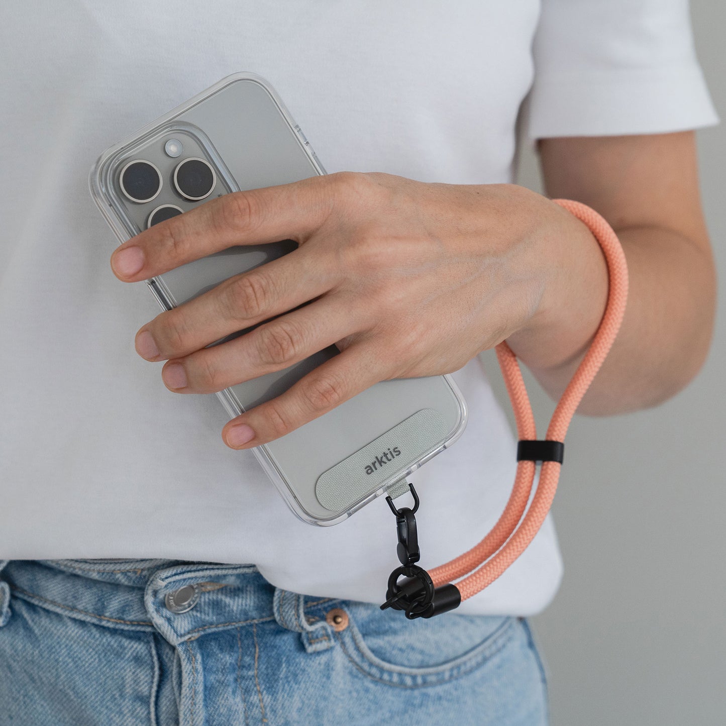 Eine Person hält ein Smartphone mit einer durchsichtigen Hülle und der pfirsichfarbenen Arktis Lanyard Handykette fürs Handgelenk von Arktis und kombiniert dieses stylische Mode-Accessoire mit Jeans und einem weißen Hemd für praktisches Smartphone-Zubehör und modernes Flair.