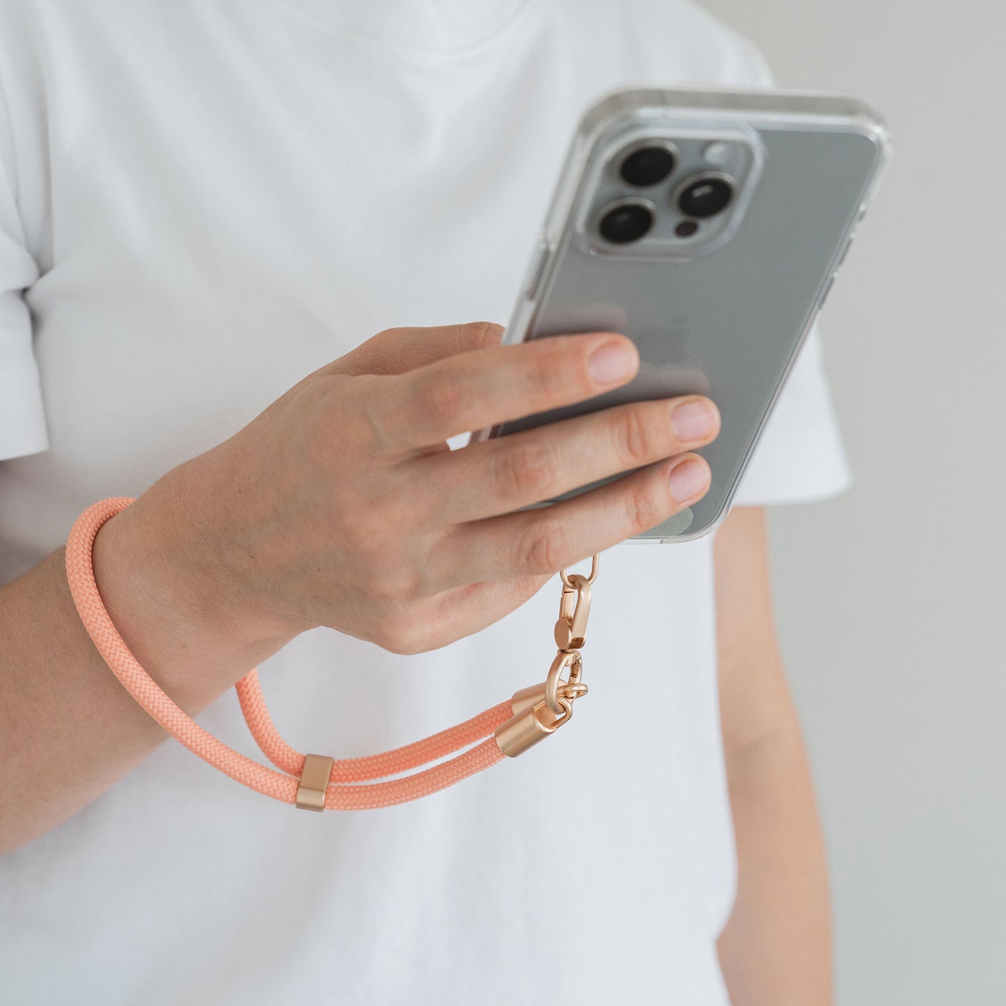 Eine Person in einem weißen T-Shirt hält ein Smartphone mit der Arktis Lanyard Handykette fürs Handgelenk von Arktis. Ein orangefarbenes Armband mit goldenen Beschlägen ist um ihr Handgelenk geschlungen. Der Hintergrund ist schlicht und hell.