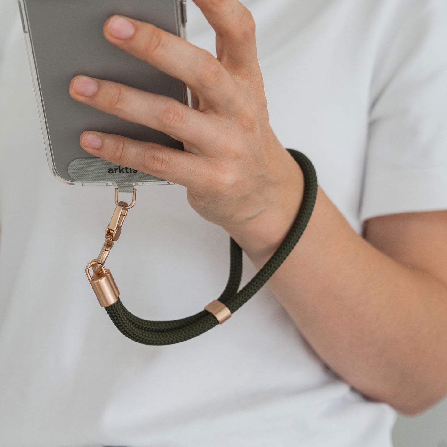 Eine Person im weißen Hemd hält ein Smartphone, an dem die arktis Lanyard Handykette fürs Handgelenk von Arktis in Grün und Gold befestigt ist. Ihre Hand und ihr Arm heben dieses stylische Smartphone-Accessoire hervor.