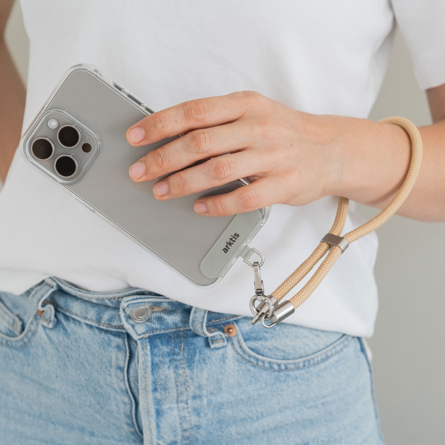Eine Hand hält ein Smartphone mit der arktis Lanyard Handykette fürs Handgelenk von Arktis in einer durchsichtigen Hülle, getragen mit Jeans und weißem Hemd – ideales Smartphone-Zubehör für modebewusste Benutzer aus der Mode-Accessoire-Linie von Arktis.