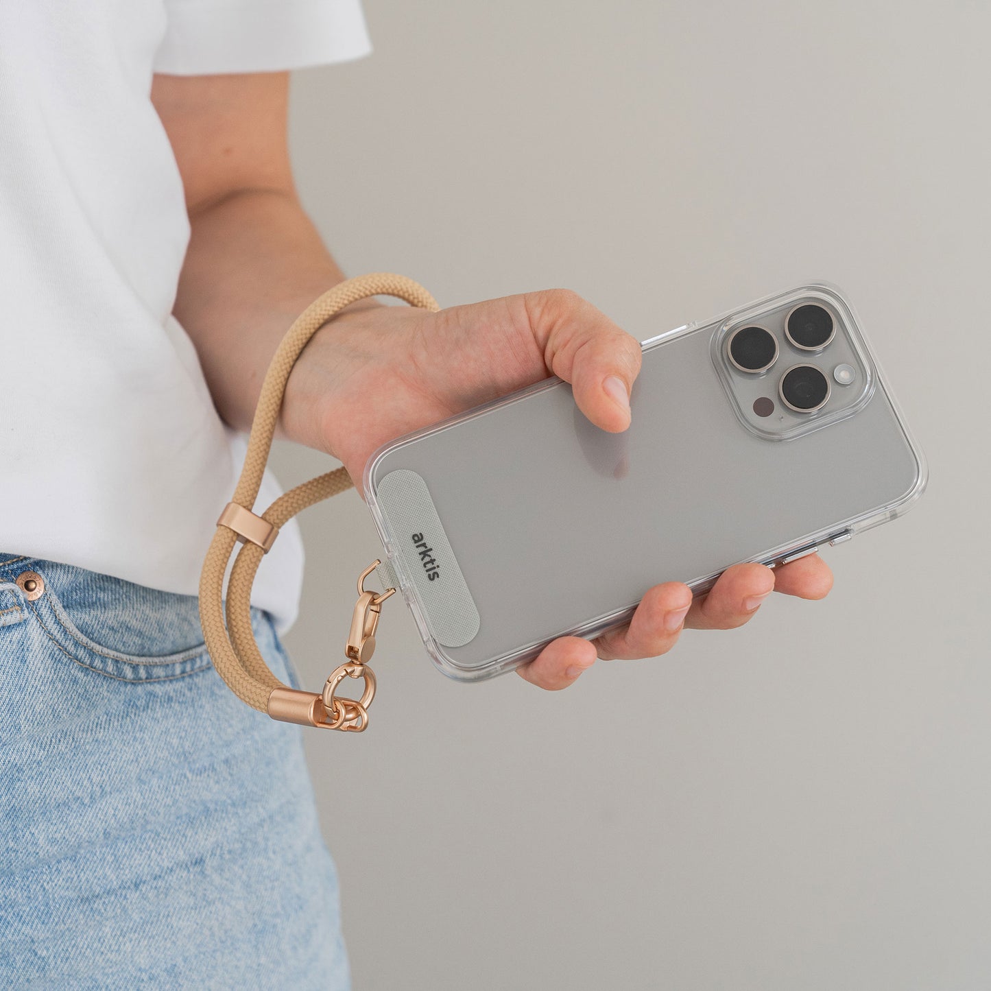 Eine Person in hellblauer Jeans und weißem Hemd hält ein Smartphone mit der Arktis Lanyard Handykette fürs Handgelenk von Arktis. Die Handykette verfügt über ein transparentes Gehäuse und ein beiges Armband mit goldenen Beschlägen. Der Hintergrund ist schlicht hellgrau.