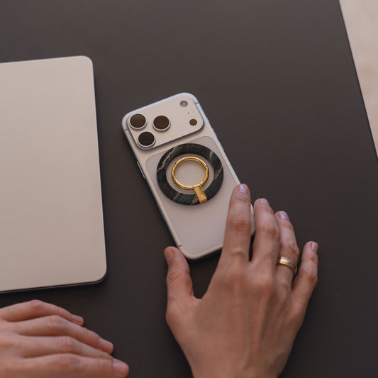 Eine Hand mit einem Goldring ruht neben einem arktis Magnetischer Ringhalter für MagSafe - Sweet Leopard, abgebildet mit iPhone Zubehör und einem geschlossenen Laptop auf einer dunklen Fläche.