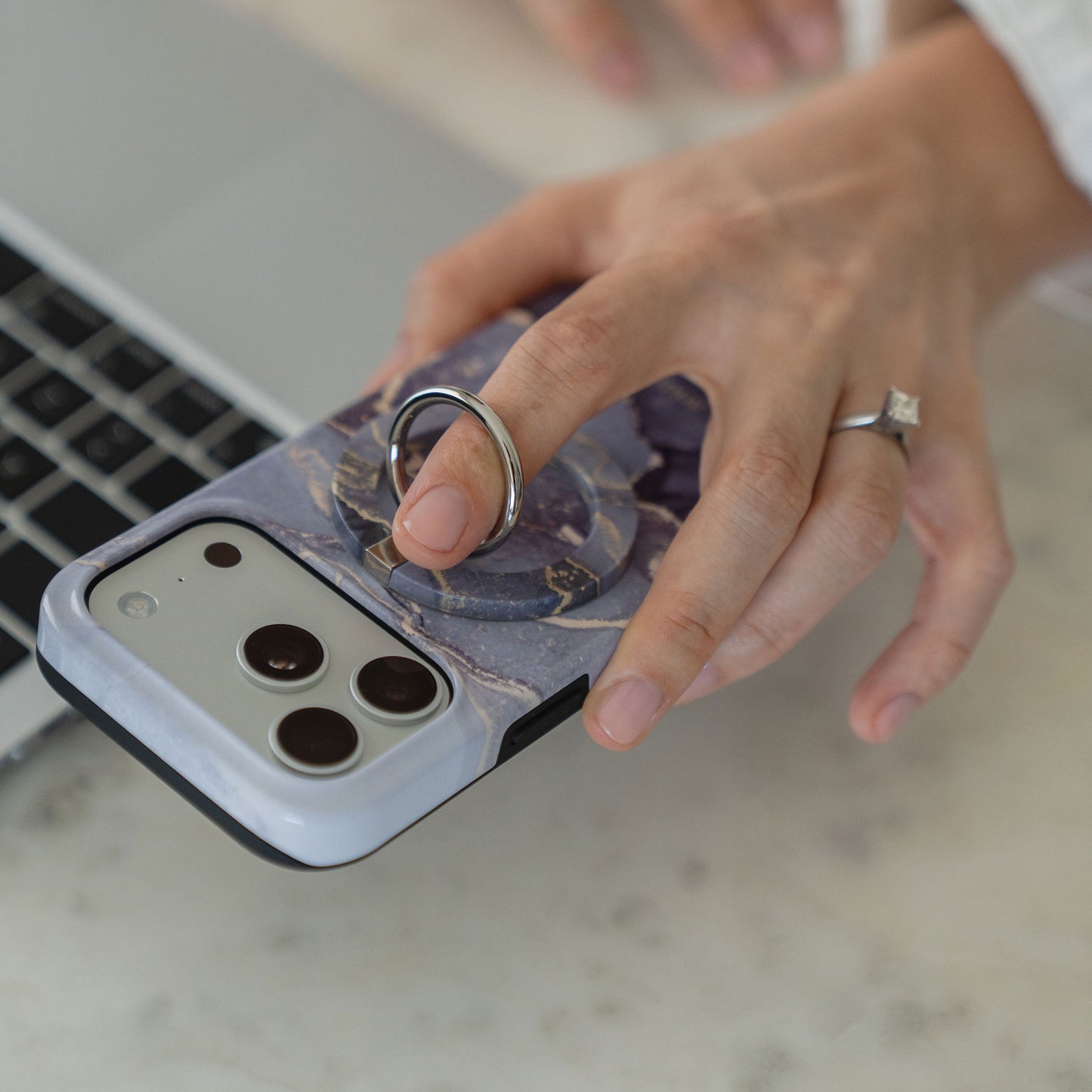 Eine Hand mit einem Ring hält die arktis iPhone 14 Pro Max Hülle - Dusty Marble, eine MagSafe-kompatible Hülle mit Ringhalterung, neben einem Laptop auf einer hellen Oberfläche.