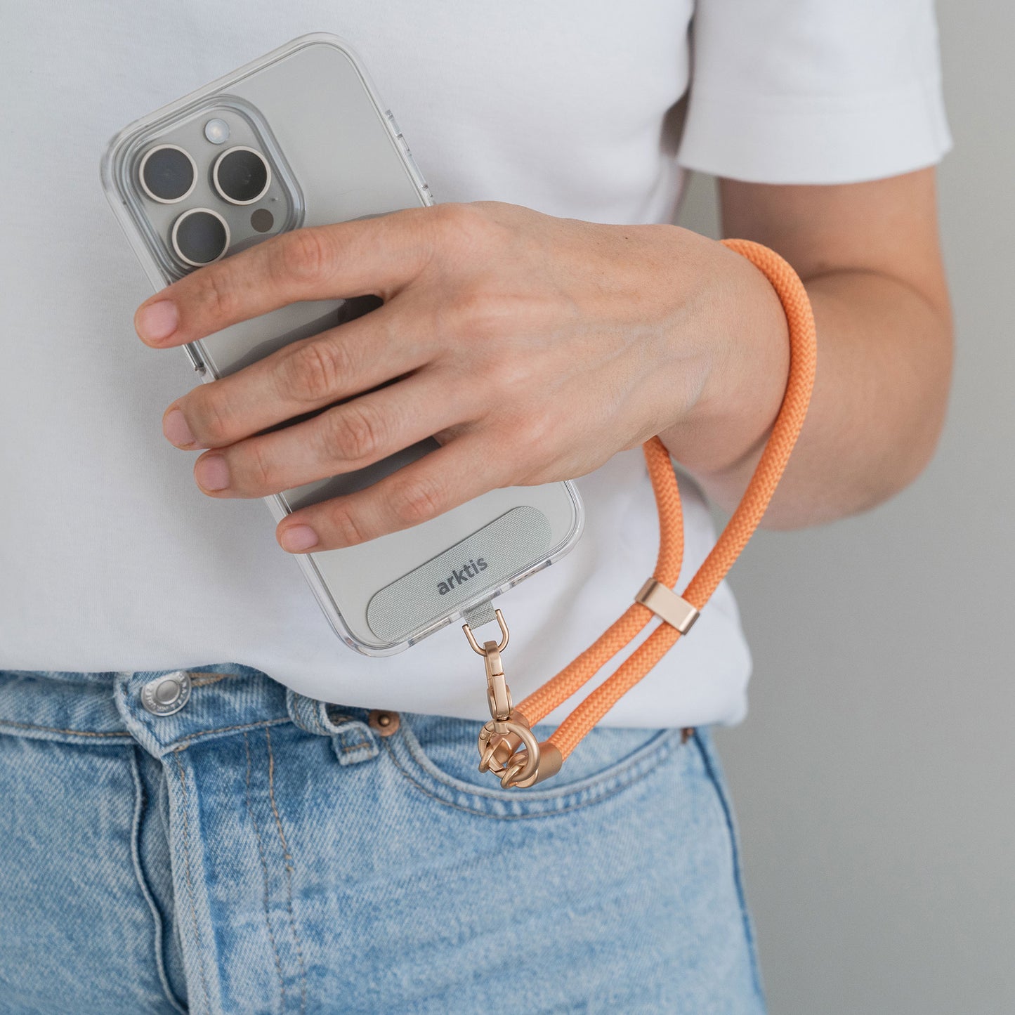 Eine Person in einem weißen Hemd und blauen Jeans hält ein Smartphone mit einer durchsichtigen Hülle, die mit der Arktis Lanyard Handykette fürs Handgelenk in Orange versehen ist, was sie zu einem stilvollen und praktischen Accessoire macht.