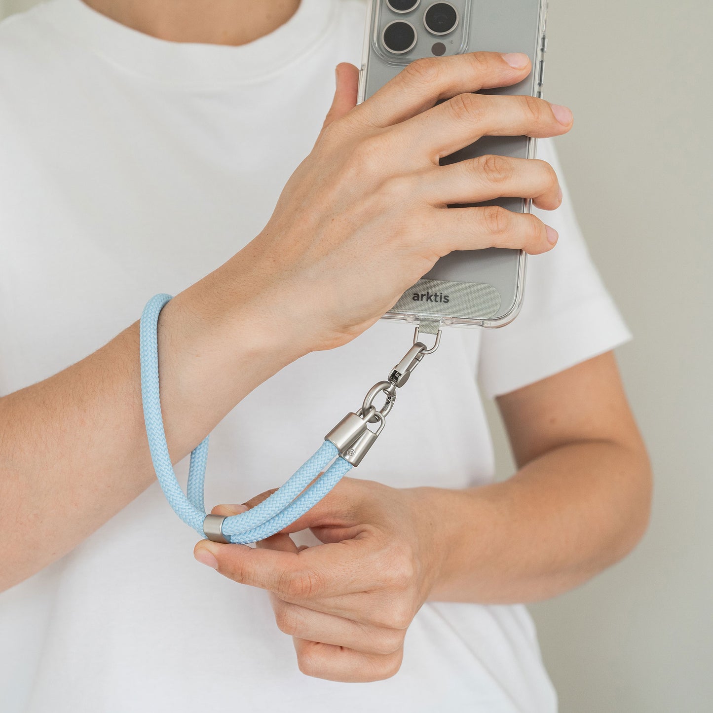 Eine Person in einem weißen Hemd hält ein Telefon, an dem die arktis Lanyard Handykette fürs Handgelenk von Arktis befestigt ist – eine Kombination aus Smartphone-Zubehör und stilvollem Mode-Accessoire.