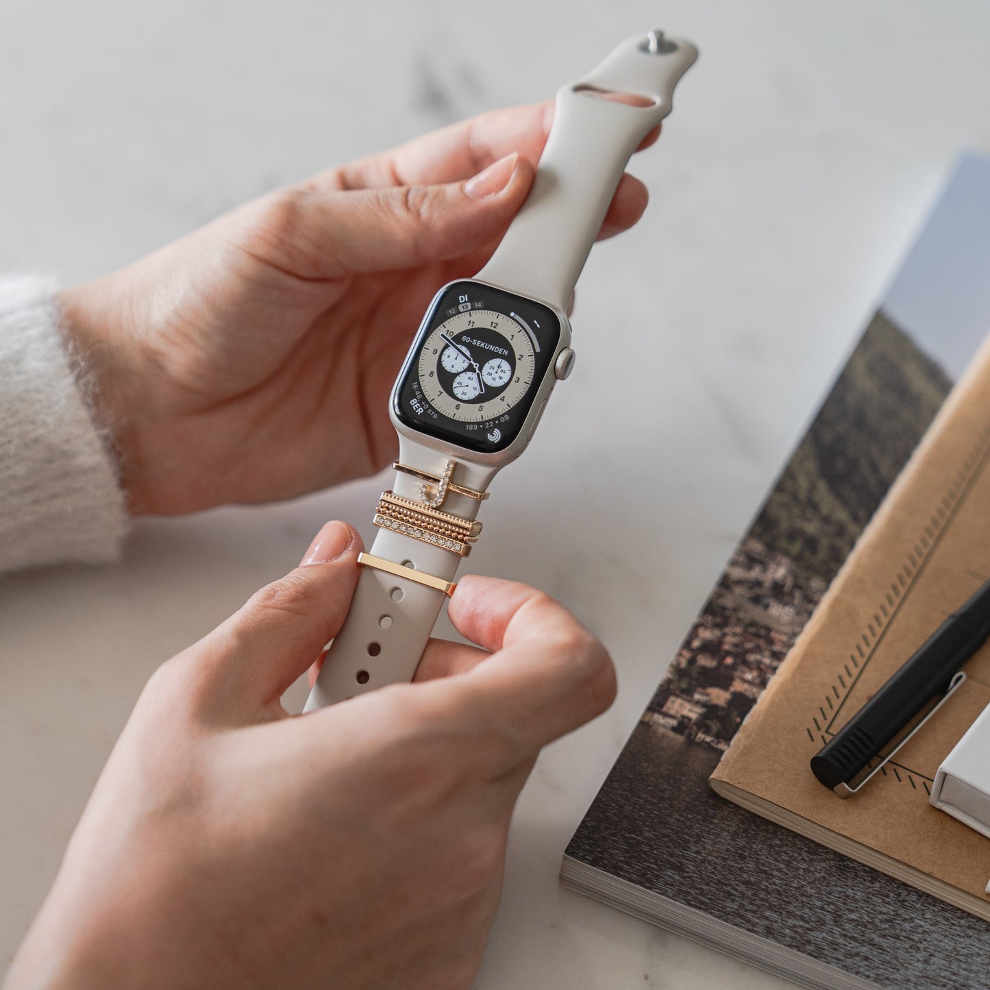 Eine Person hält eine Arktisband Apple Watch Charms „J Style“-Smartwatch mit beigem Arktisband, das für einen extravaganten Look sorgt, neben Notizbüchern und einem Stift auf dem Tisch.