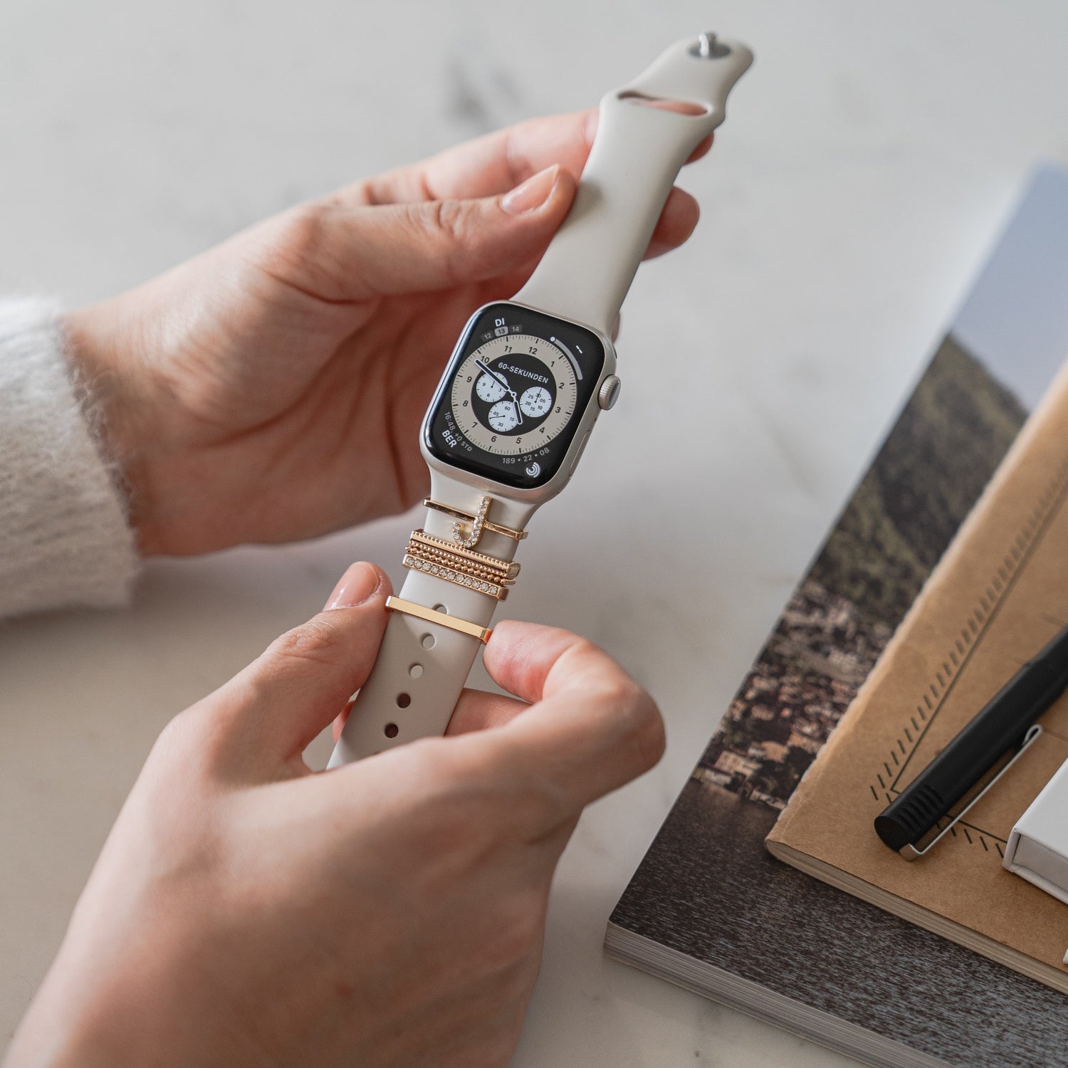 Eine Person hält eine Arktisband Apple Watch Charms „J Style“-Smartwatch mit beigem Arktisband, das für einen extravaganten Look sorgt, neben Notizbüchern und einem Stift auf dem Tisch.