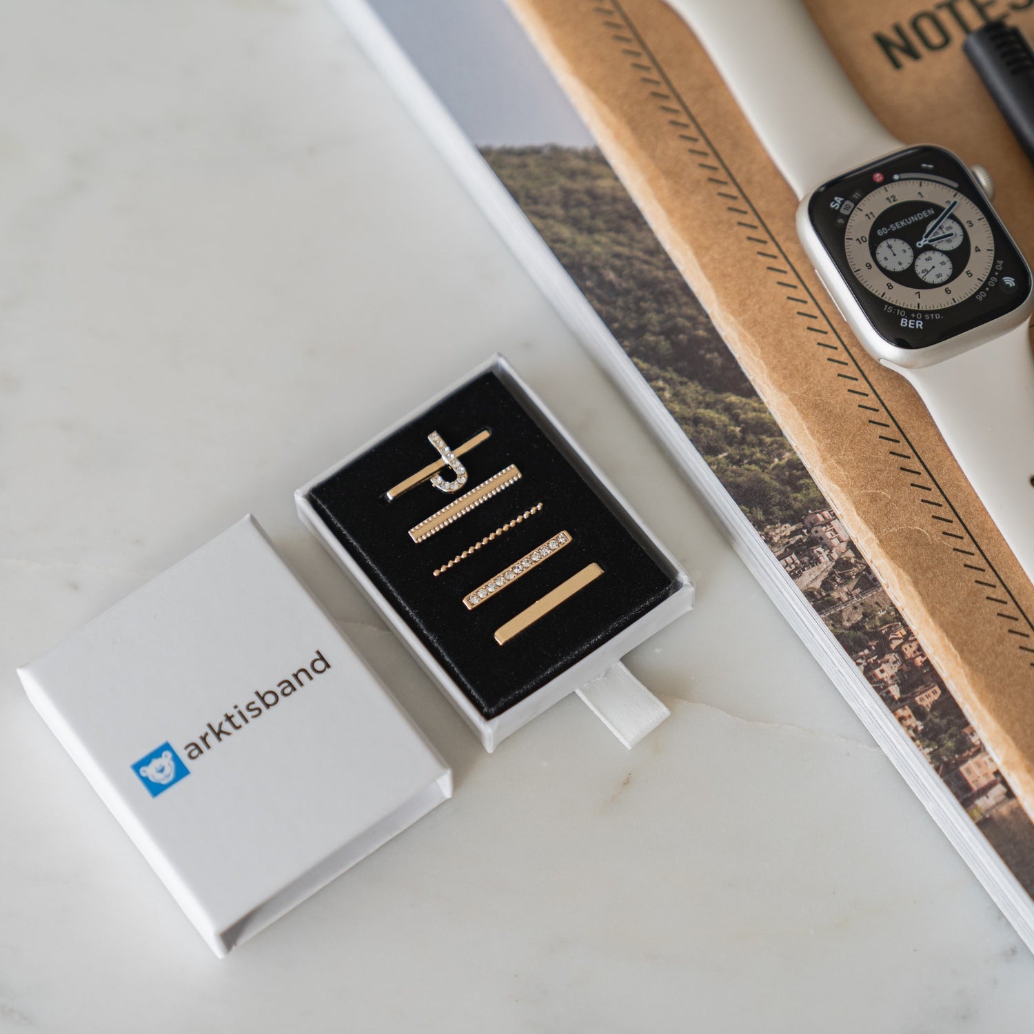 Eine weiße Box mit der Aufschrift „arktisband“ steht neben goldenem Uhrenzubehör, einer Smartwatch mit Arktisband Apple Watch Charms „J Style“ und Notizbüchern auf einem Tisch und sorgt für einen extravaganten Look.