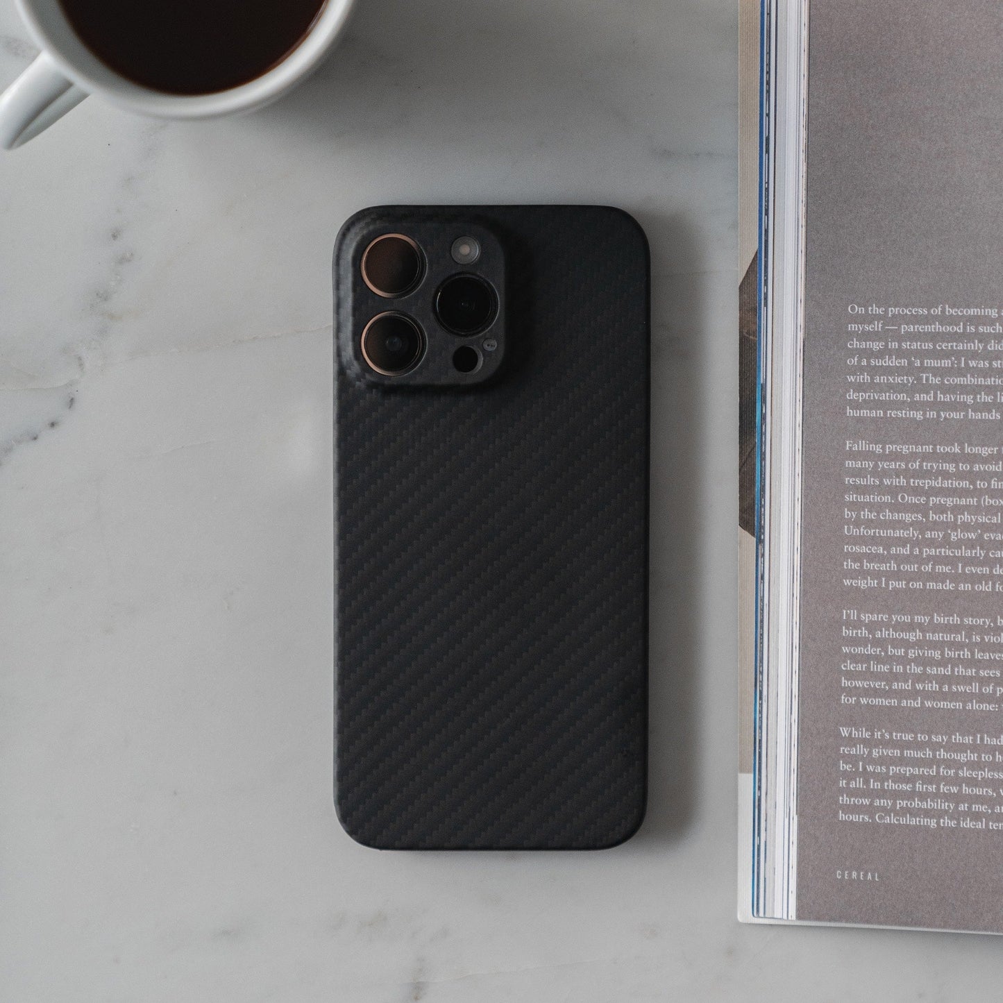 Ein arktis iPhone 16 Pro Carbon Case aus Carbon- und Aramidfasern schützt ein Smartphone, das neben einer offenen Zeitschrift und einer Kaffeetasse auf Marmor liegt.