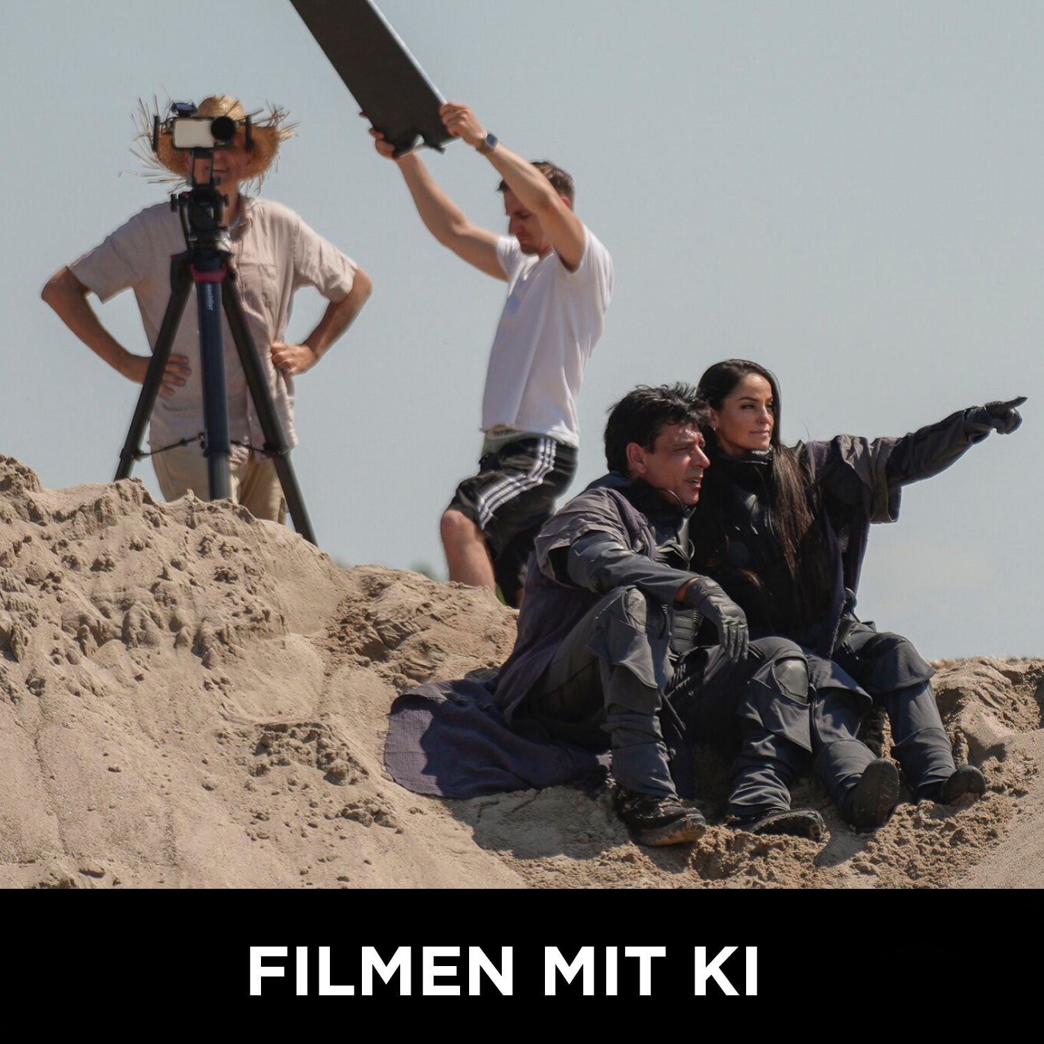 Zwei kostümierte Personen sitzen im Sand, eine zeigt nach vorne; dahinter hält eine Person einen Reflektor, eine andere steht neben einer Kamera. „FILMEN MIT KI“ erscheint unten. Lerne Filmemachen mit Rainer Wolfs Wolffilms Academy Online-Kurs – Einfach filmen lernen.