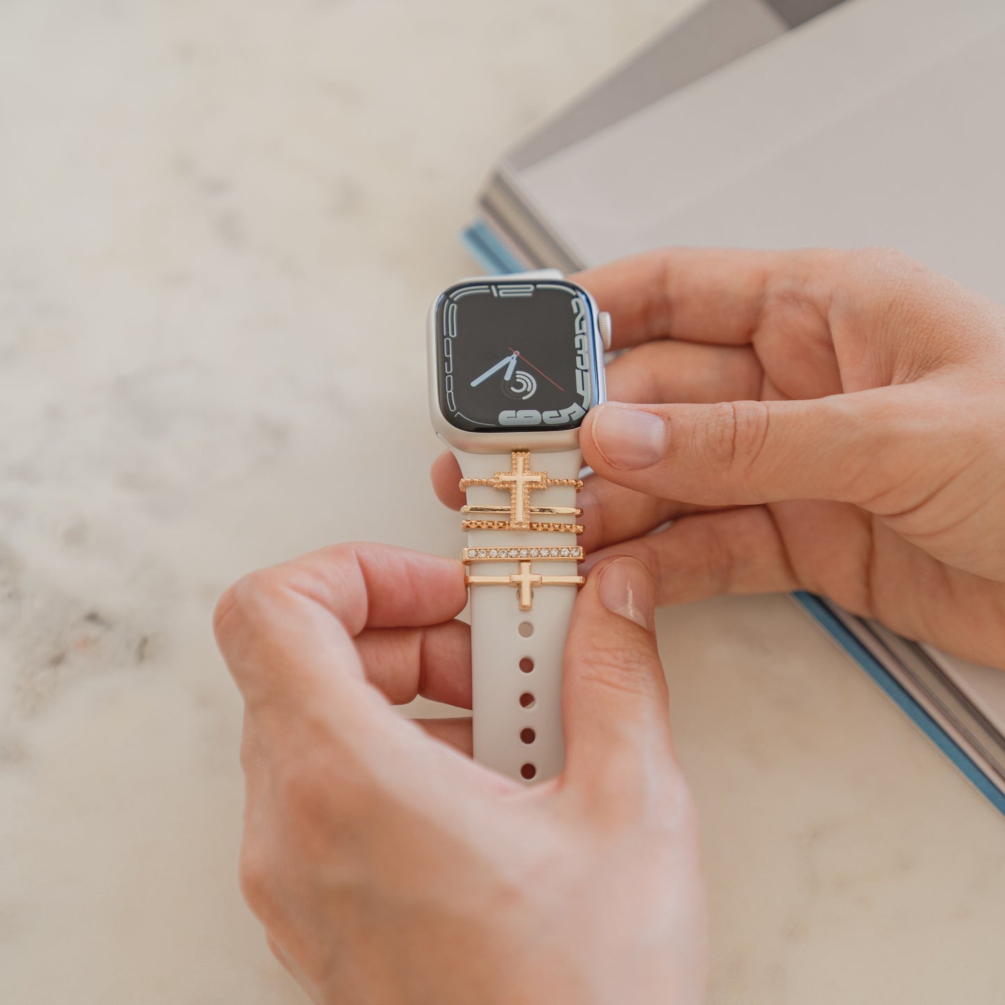 Zeiger halten eine Smartwatch mit beigem Arktisband-Armband, verziert mit Arktisband-Apple-Watch-Charms „Kreuz“ in Gold.