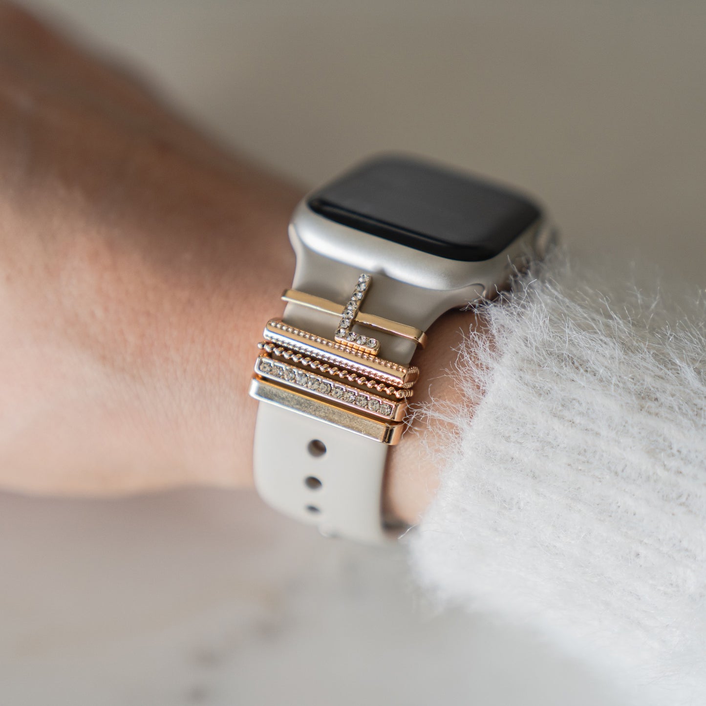 Eine Nahaufnahme eines Handgelenks, das ein Arktisband Apple Watch Armband mit „L Style“-Anhängern trägt, mit übereinander gestapelten Gold- und Silberringen – einige davon mit kleinen Diamanten verziert. Die Person trägt einen flauschigen weißen Pullover.