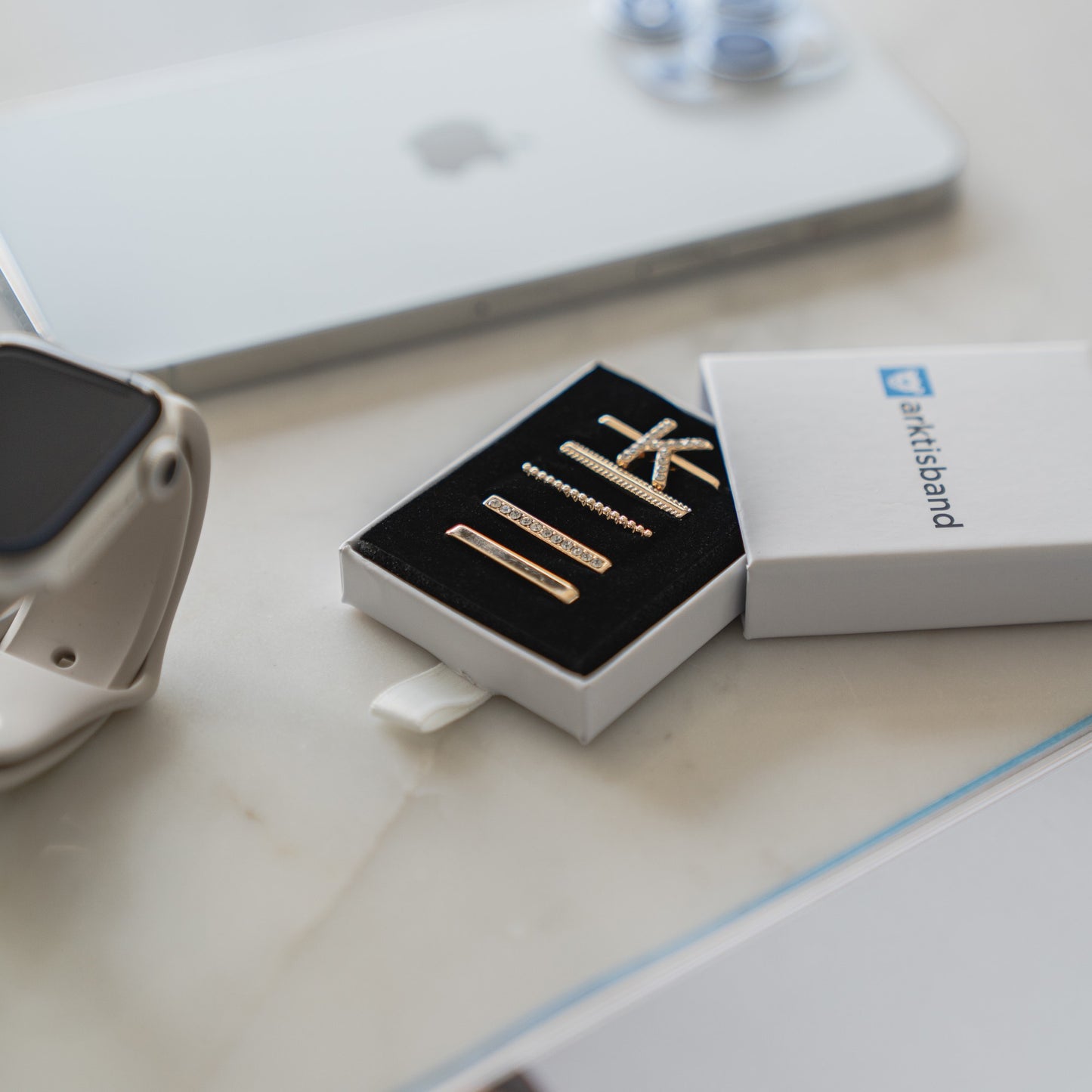 Eine Schachtel mit metallischen Krawattenklammern auf einem Schreibtisch neben einem Smartphone und einer Apple Watch, verziert mit Arktisband Apple Watch Charms „K Style“ von Arktisband.
