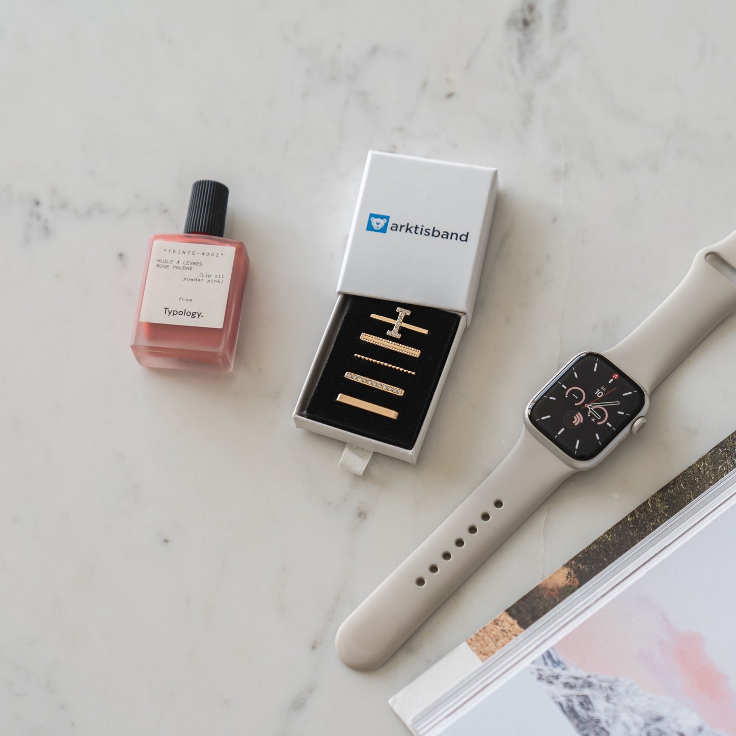 Eine Flasche Typology-Parfüm, eine offene Arktisband-Box mit vier Goldbarren und T-förmigen Edelstahlanhängern, ein Arktisband „I Style“-Apple-Watch-Anhänger in Beige und ein Teil eines Buches liegen auf einer weißen Marmoroberfläche.