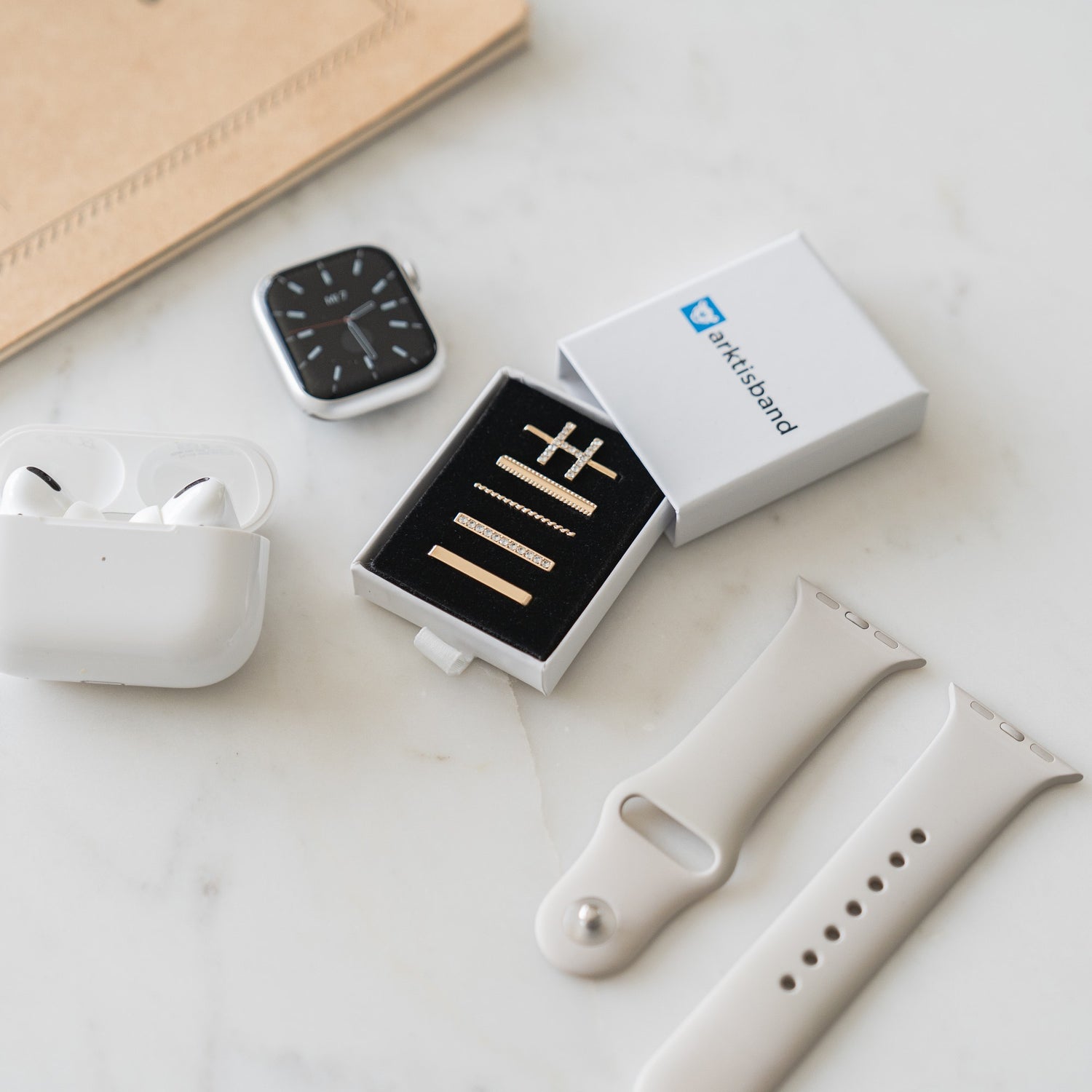 Eine Smartwatch, Ohrhörer und beige Arktisband-Uhrenarmbänder mit Arktisband-Apple-Watch-Charms „H Style“ liegen auf einer weißen Oberfläche neben einer kleinen Box mit stilvollen goldenen Accessoires.