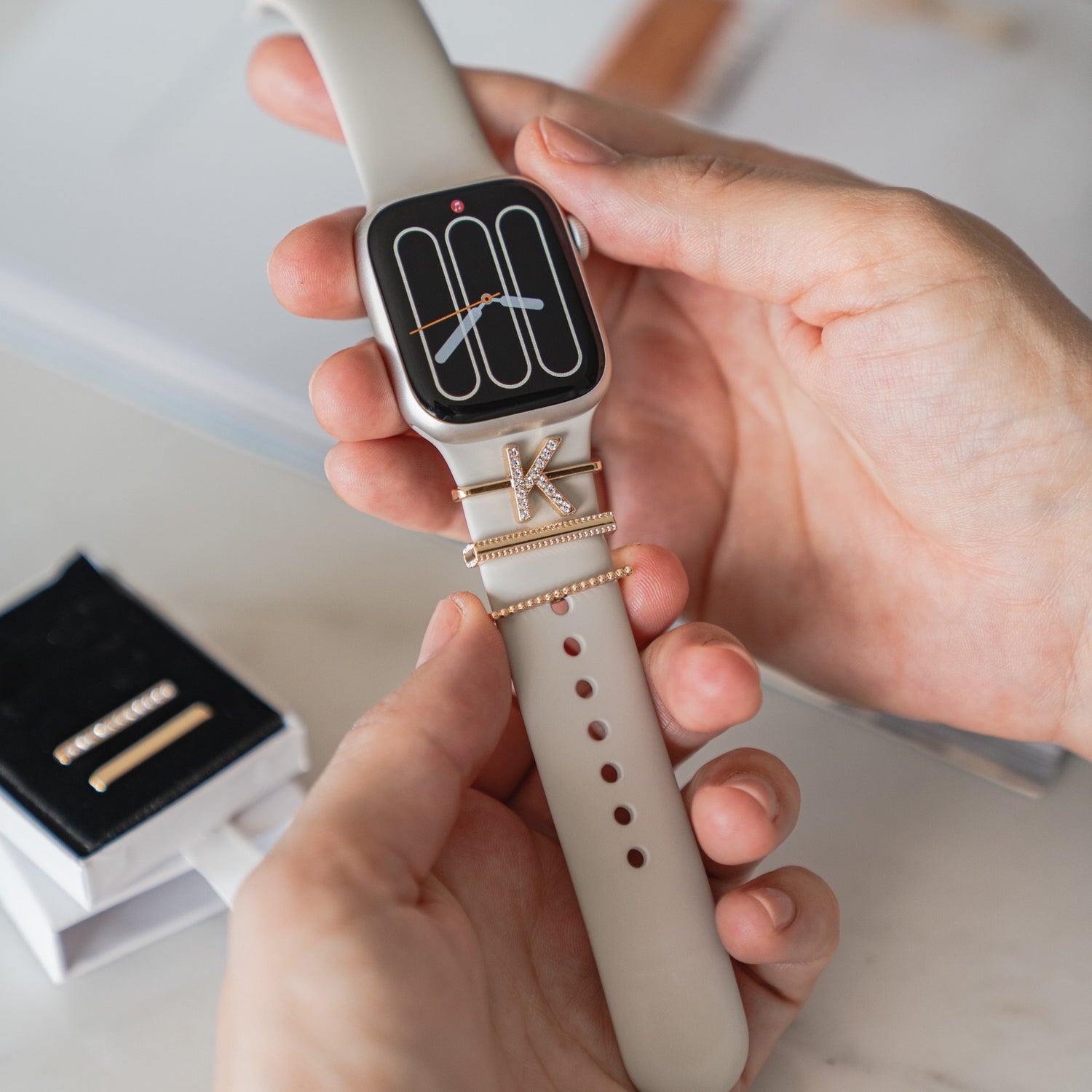 Eine Hand zeigt eine Apple Watch mit Arktisband Apple Watch Charms „K Style“ auf ihrem beigen Arktisband-Armband; daneben steht eine Schmuckschatulle.