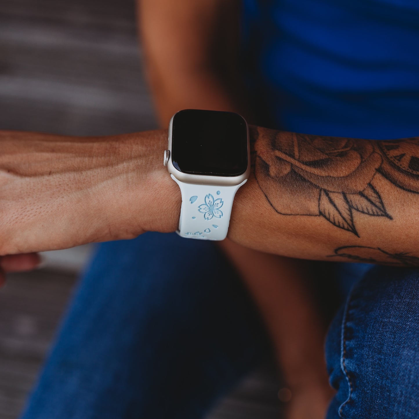 Nahaufnahme des Handgelenks einer Person mit dem arktisband Apple Watch Armband „Flores“ von arktisband, einem weißen Silikonarmband mit blauem Blumenmuster. Die Person trägt ein Blumentattoo und Blue Jeans.