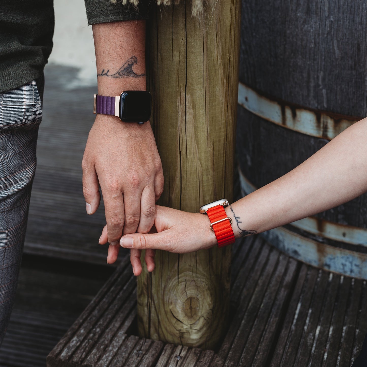 Zwei Menschen mit passenden Vogeltattoos halten sich an einem Holzpfosten an den Händen und tragen jeweils eine Apple Watch. Einer trägt ein Arktisband Kunstlederarmband „Gustino“ in Lila mit Magnetverschluss, der andere ein rotes Armband.