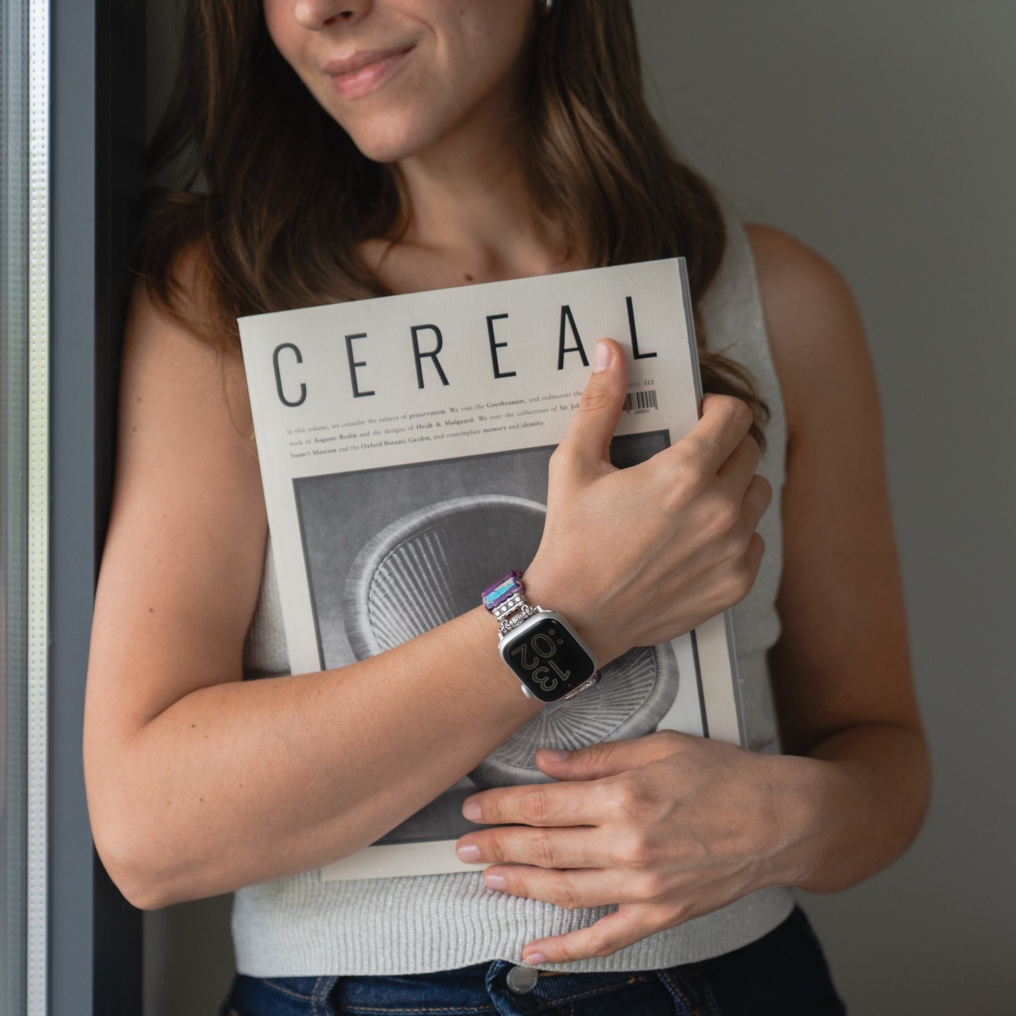 Frau im ärmellosen Oberteil, die eine CEREAL-Zeitschrift in der Hand hält, trägt die arktisband Apple Watch "Stone Loop" von arktisband und steht an einem Fenster.