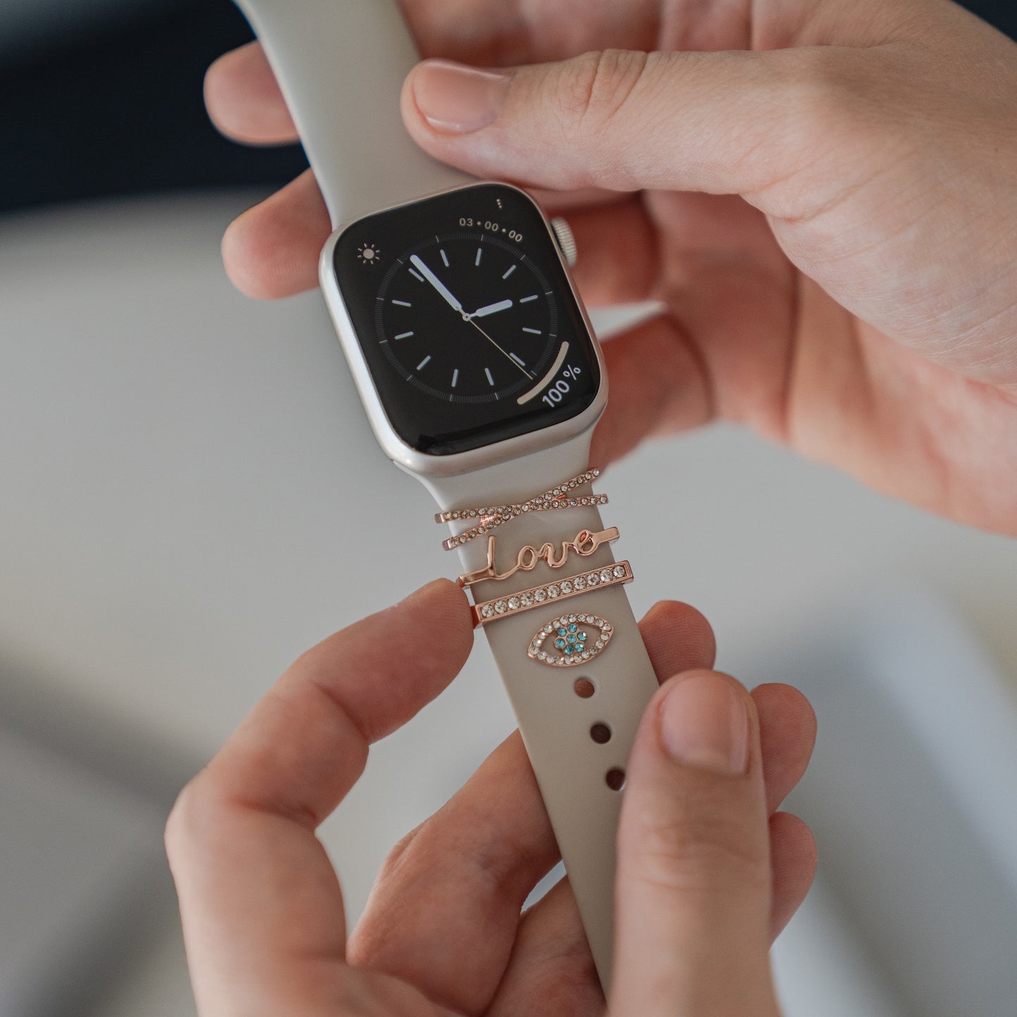 Eine Person hält eine Apple Watch mit einem hellgrauen Arktisband, geschmückt mit anpassbaren Arktisband-Apple-Watch-Armbandanhängern in Gold und mit Juwelendesigns, darunter „Love“ und ein augenförmiger Anhänger, perfekt für den persönlichen Stil.