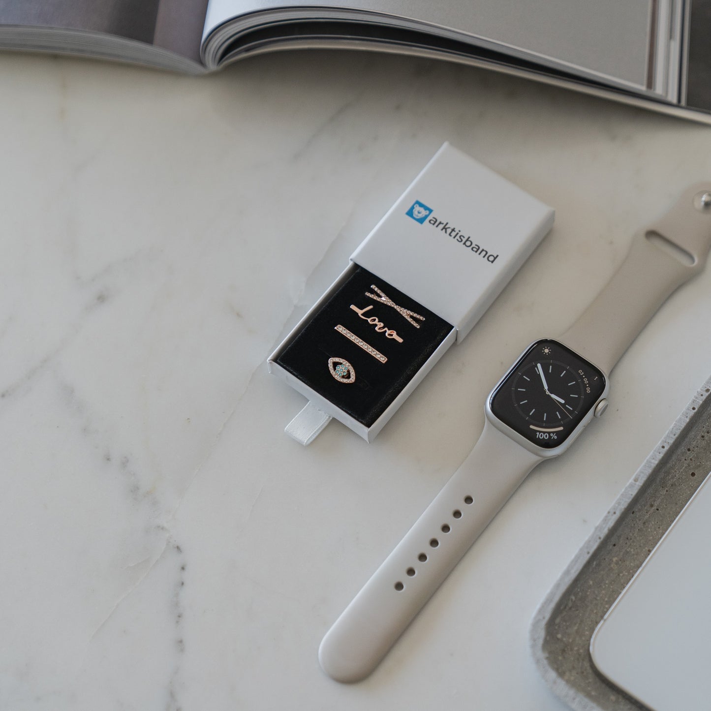 Eine Arktisband Apple Watch mit Arktisband Apple Watch Armband Charms liegt auf Marmor neben einer teilweise geöffneten weißen Schachtel, die einen Ring und eine schwarze Karte enthält. Im Hintergrund trägt eine aufgeschlagene Zeitschrift zur eleganten Szene bei.