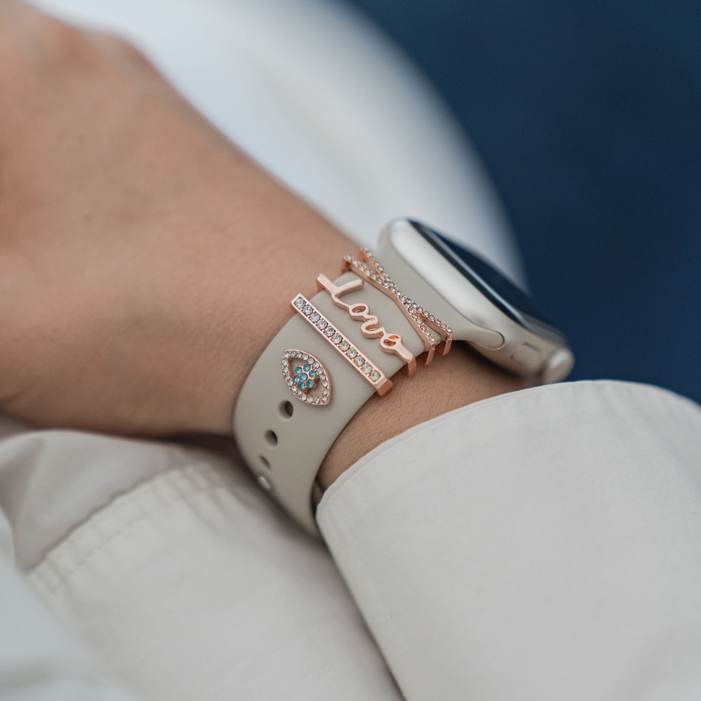 Eine Nahaufnahme zeigt ein Handgelenk mit einem arktisband Apple Watch Armband-Anhänger, der einen roségoldenen „Love“-Anhänger und einen juwelenbesetzten Anhänger in Augenform trägt. Die Trägerin, bekleidet mit einem weißen Langarmshirt, präsentiert dieses individuell gestaltete arktisband Accessoire.