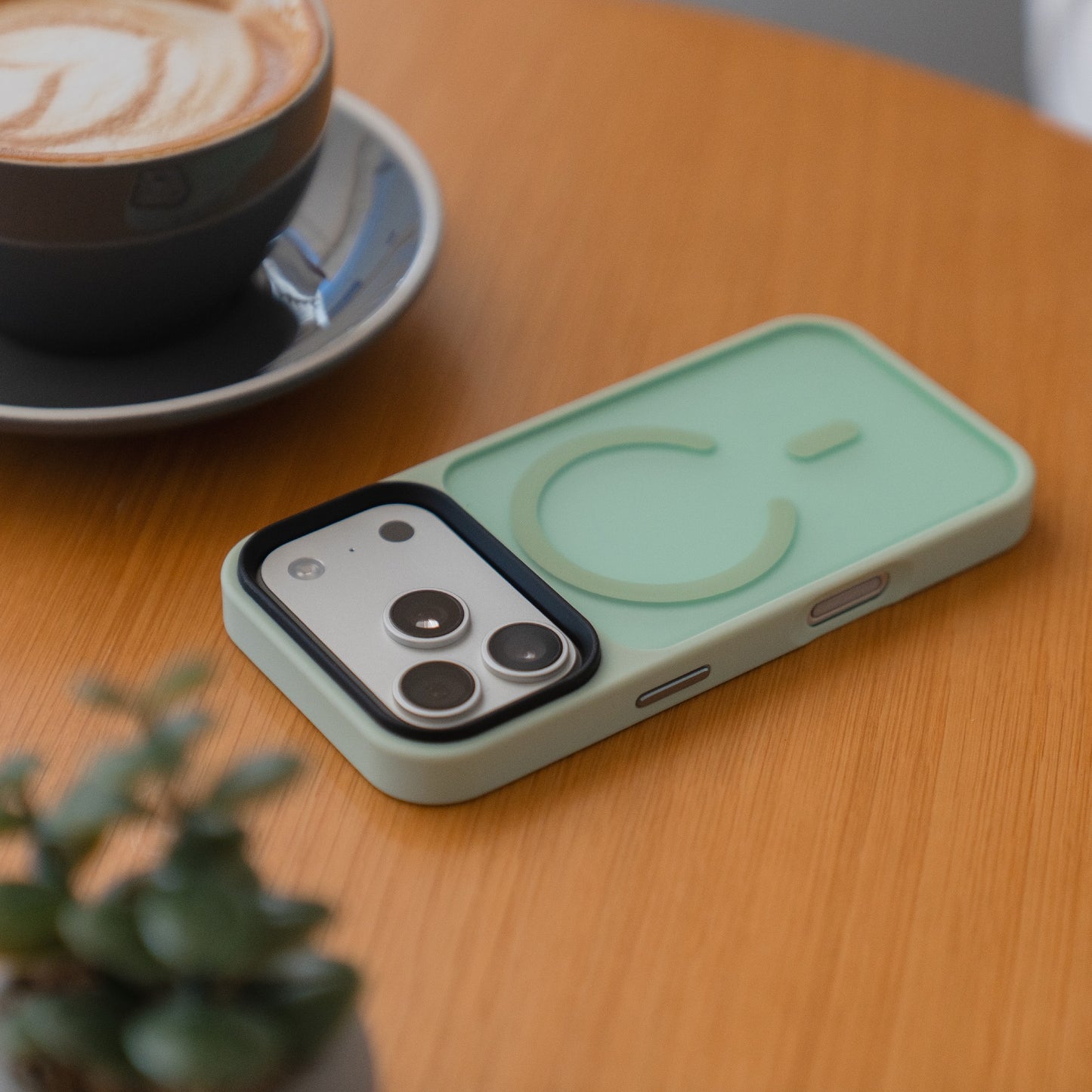 Ein iPhone 17 Pro in einem Arktis arktis Solid Case mit MagSafe in mintgrün liegt auf einem Holztisch neben einer Tasse Kaffee und einer kleinen Pflanze.