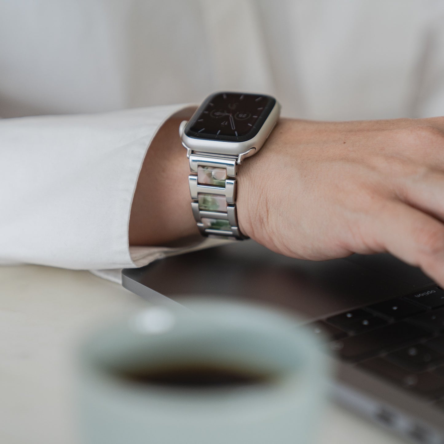 Jemand trägt ein Arktisband Apple Watch Gliederarmband „Namaste“ und tippt auf einer Laptoptastatur, im Vordergrund steht eine Tasse Kaffee auf einer weißen Fläche.