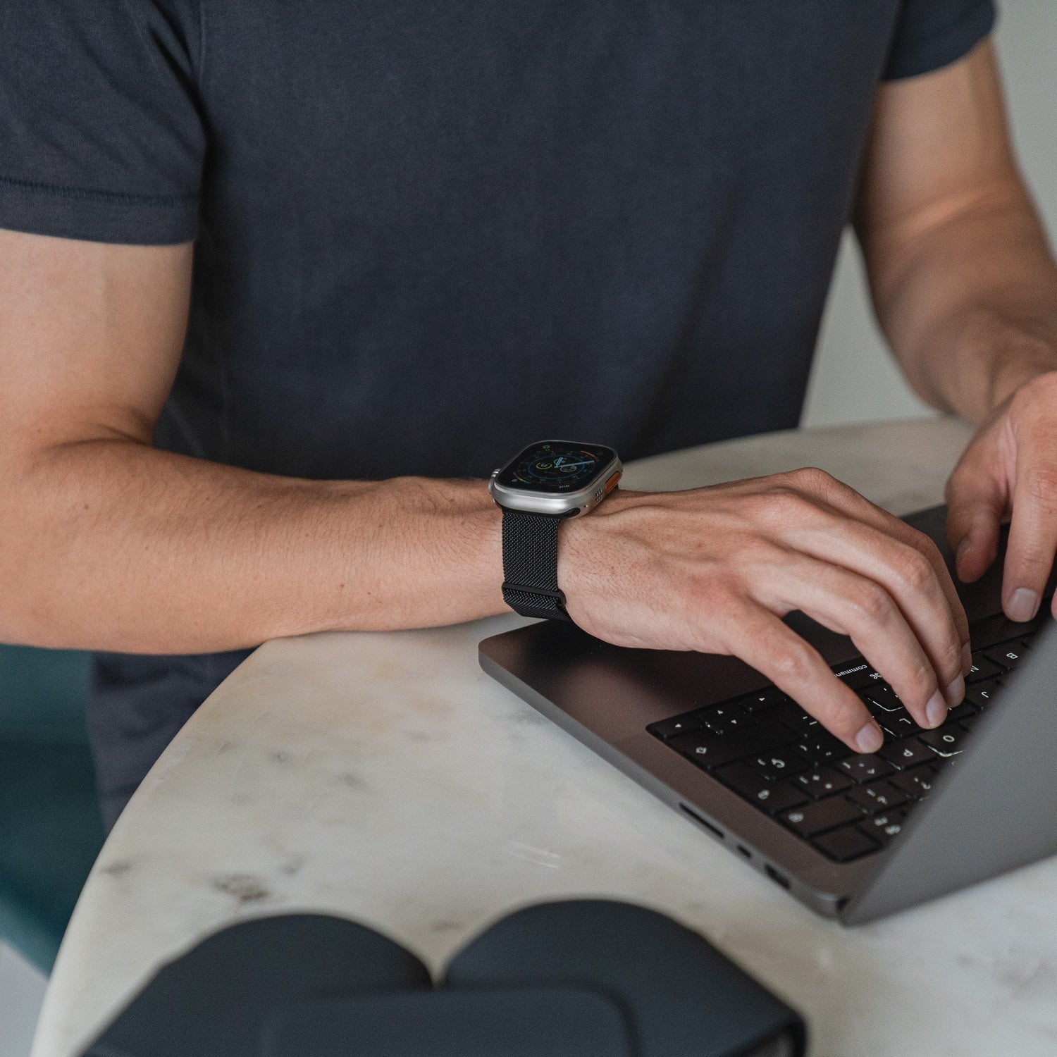 Eine Person mit einem Arktisband Apple Watch Milanaise Loop Armband tippt an einem runden Marmortisch auf einem Laptop. Im Vordergrund ist eine schwarze Tasche zu sehen, die Person trägt ein kurzärmeliges dunkles Hemd.