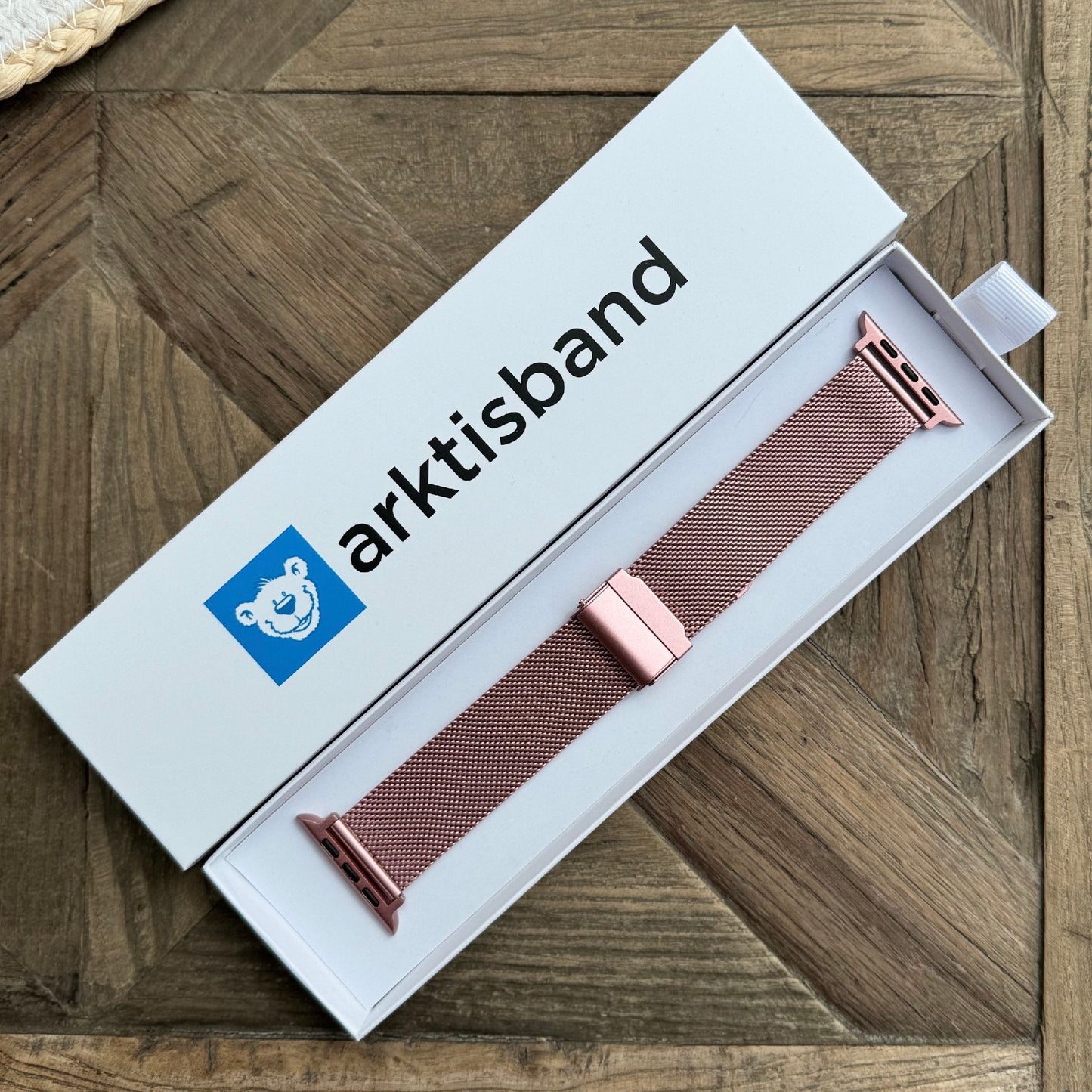 Ein Arktisband-Apple-Watch-Milanese-Armband aus rosa Edelstahlgeflecht wird in einer weißen Box mit dem Namen Arktisband und dem blauen Bärenlogo gezeigt, die auf einer Holzoberfläche ruht.