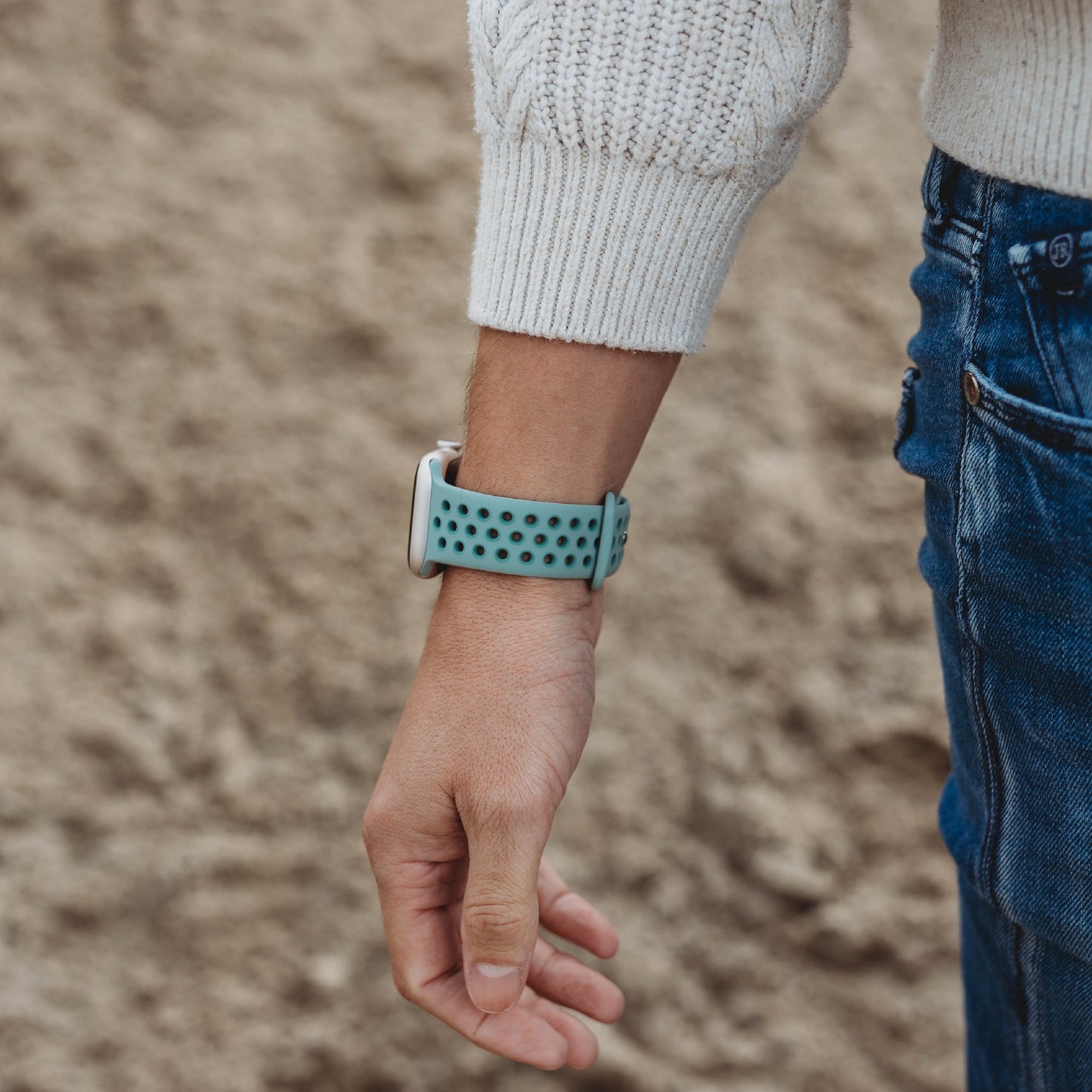 Eine Person in einem hellen Pullover und Bluejeans steht im Freien und präsentiert an ihrem linken Handgelenk ein arktisband Apple Watch Silikonarmband „Energy“ von arktisband, das ein hellgrünes Band mit runden Löchern vor einem verschwommenen braunen Hintergrund aufweist.