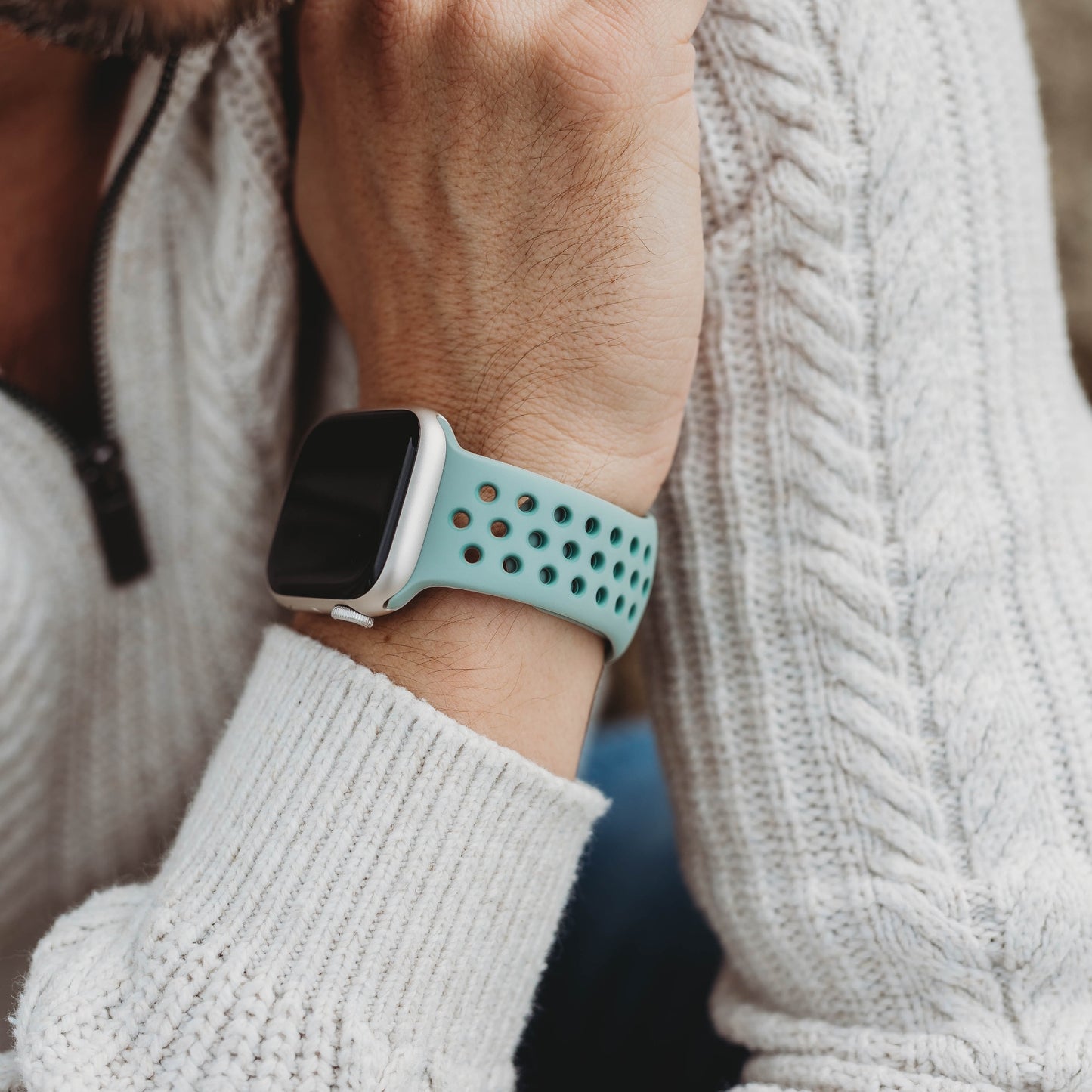 Eine Person in einem weißen Zopfmusterpullover ruht auf dem Arm und zeigt das arktisband Apple Watch Silikonarmband "Energy" in Hellblau mit schwarzen Löchern. Das Zifferblatt ist leer, ein Teil des Kinns ist sichtbar.