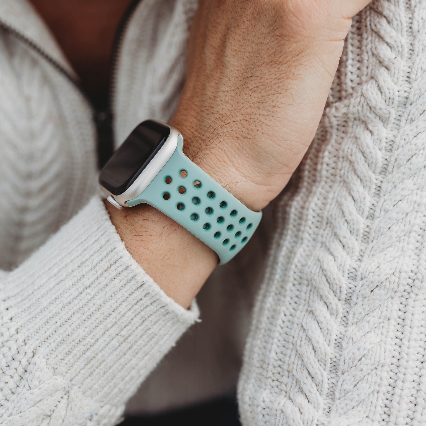Eine Nahaufnahme eines Handgelenks mit dem türkisfarbenen Arktisband Apple Watch Silikonarmband „Energy“ mit mehreren Löchern. Die Person trägt einen weißen Strickpullover mit sichtbarem Reißverschluss.