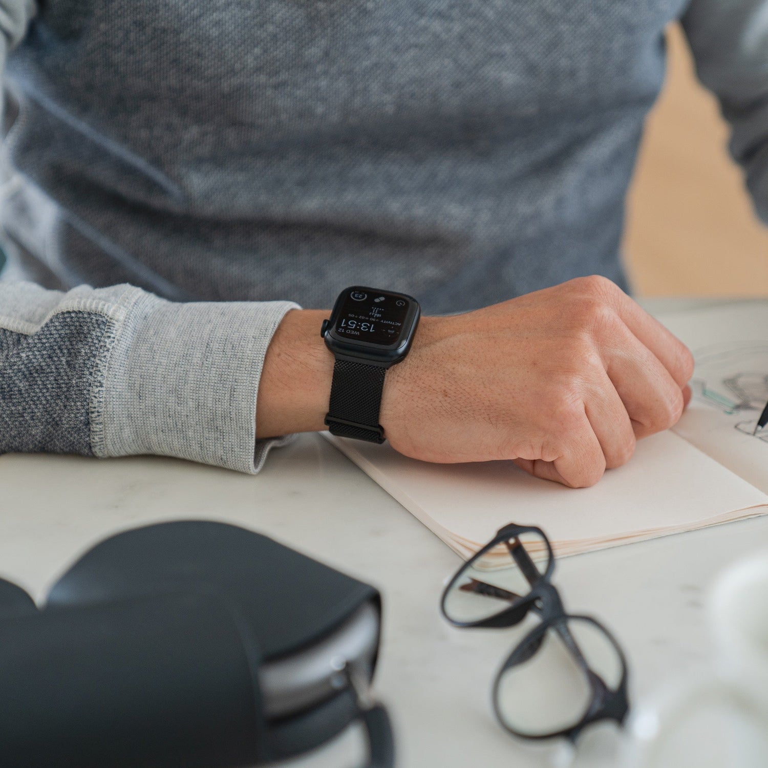 Eine Person mit einem Arktisband Apple Watch Milanaise Loop Armband sitzt an einem Tisch. In der Nähe befinden sich ein offenes Notizbuch, eine Brille und ein schwarzes Etui. Sie trägt ein graues Langarmhemd.
