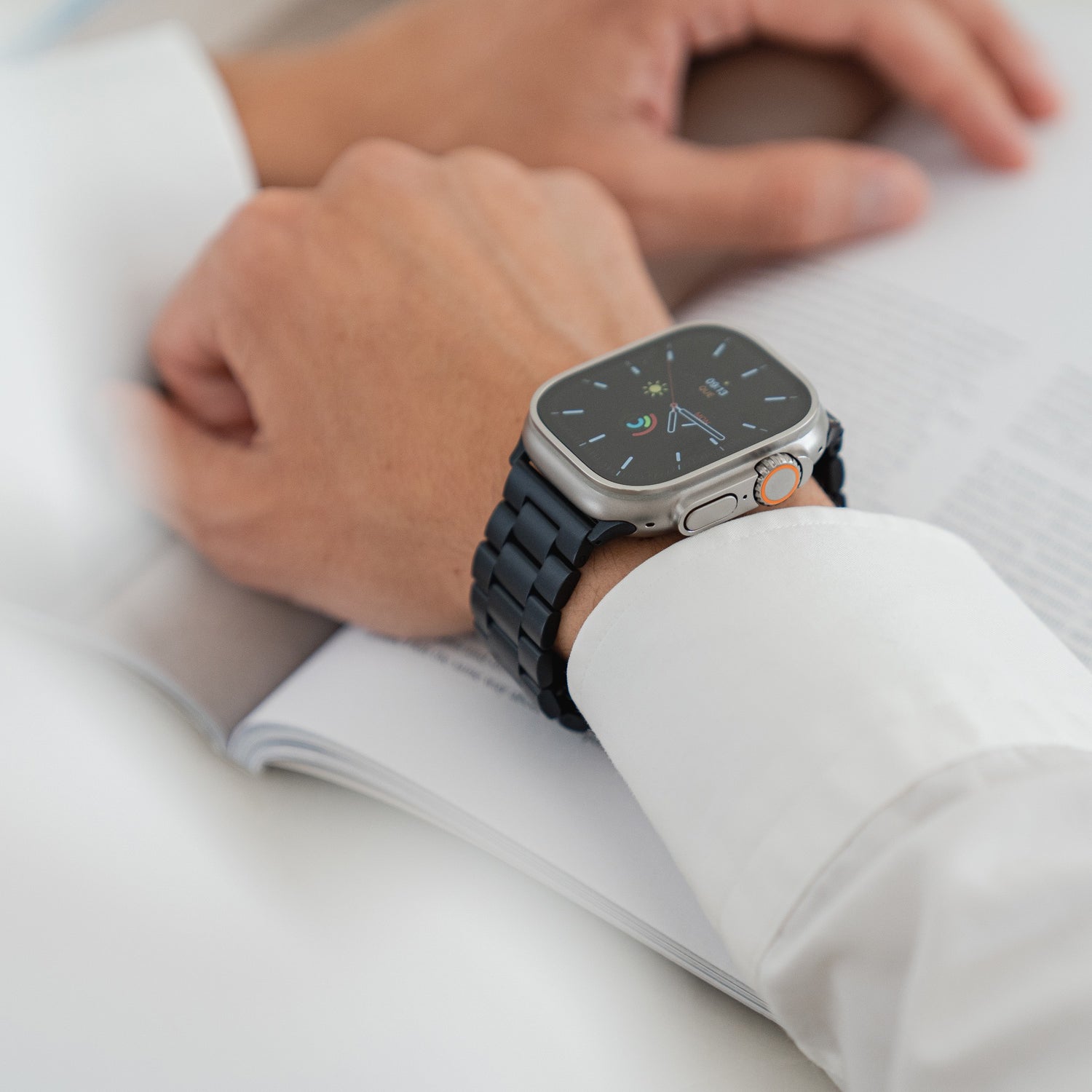 Eine Person im weißen Langarmshirt trägt das Arktisband Apple Watch Gliederarmband „Arctica“ und legt ihre Hände auf ein aufgeschlagenes Buch oder eine Zeitschrift.