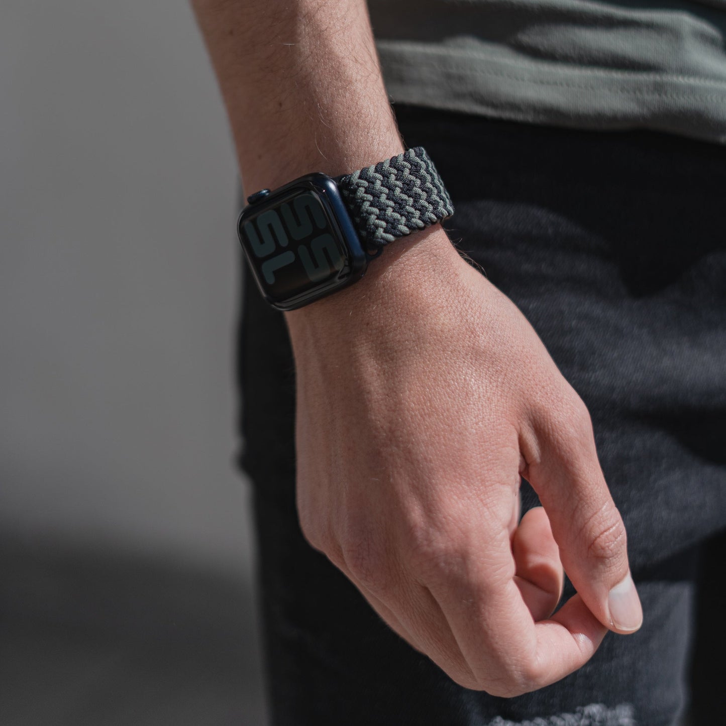 Eine Nahaufnahme der linken Hand und des Handgelenks einer Person, die das geflochtene Flex Loop TwoTone Armband mit Edelstahlverschluss der Apple Watch trägt. Die Person trägt ein graues Hemd und dunkle Jeans und steht im Tageslicht.