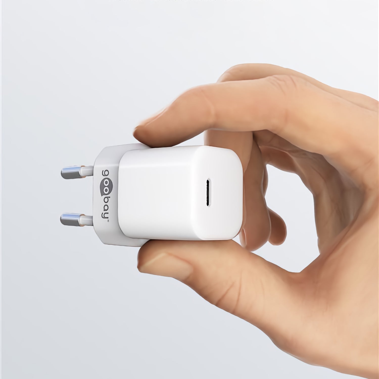 Eine Hand hält vor einem schlichten Hintergrund ein weißes Goobay USB-C PD GaN Schnellladegerät Nano 20 W-Wandladegerät mit zwei runden Stiften und Power Delivery 3.0 für schnelles Laden.