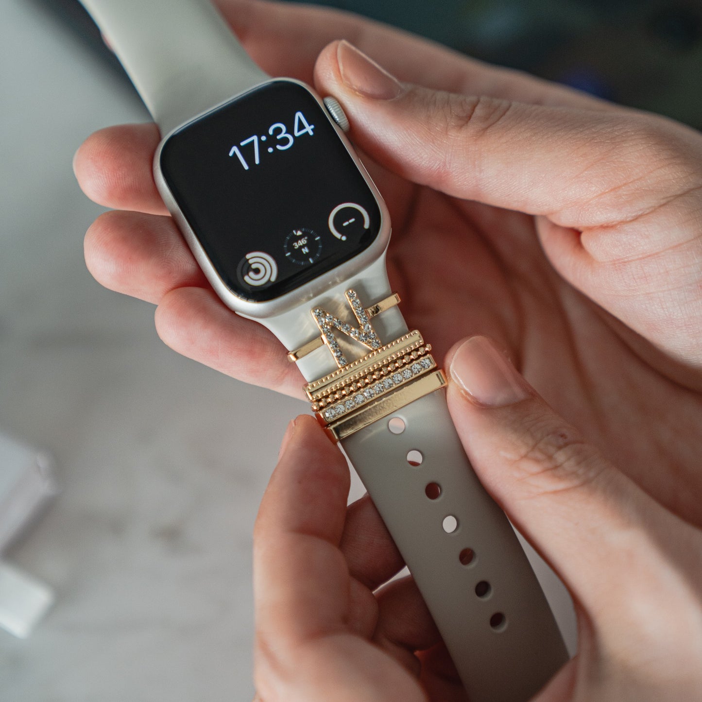 Eine Person befestigt Arktisband Apple Watch Charms „N Style“ und funkelnde Edelstahl-Charms an einem hellen Arktisband-Armband ihrer Smartwatch, die die Uhrzeit 17:34 anzeigt.