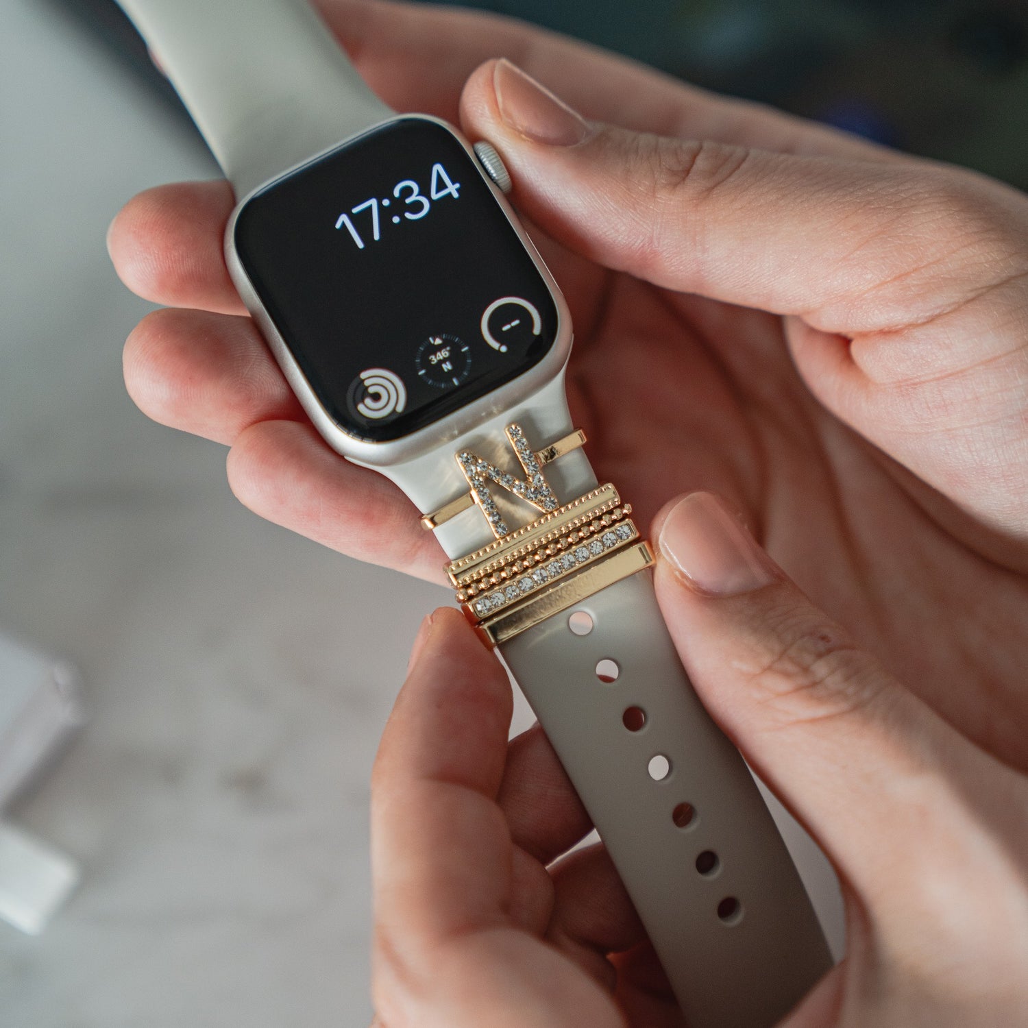 Eine Person befestigt Arktisband Apple Watch Charms „N Style“ und funkelnde Edelstahl-Charms an einem hellen Arktisband-Armband ihrer Smartwatch, die die Uhrzeit 17:34 anzeigt.