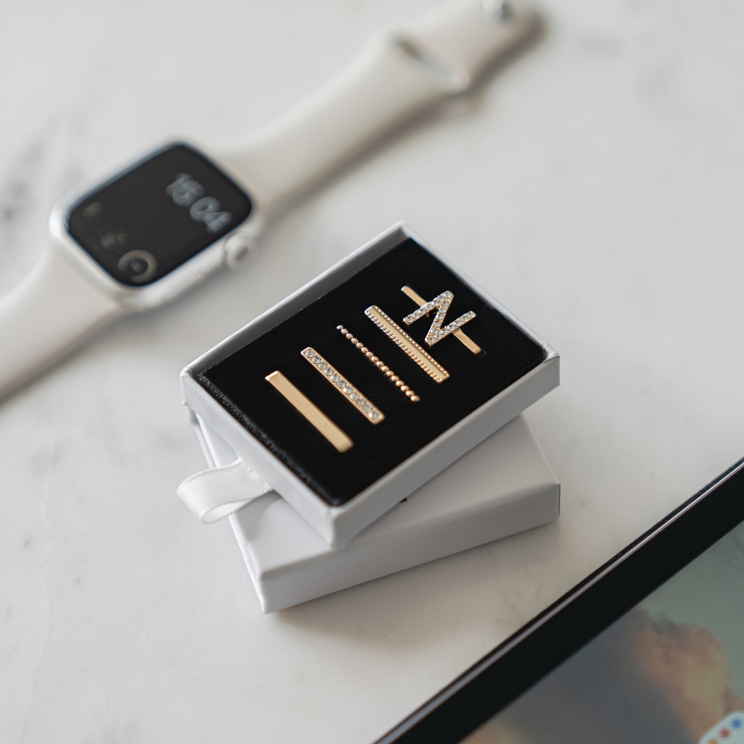 Ein Satz goldener Ohrstecker in einer Schachtel liegt neben einer weißen Smartwatch, die mit Arktisband Apple Watch Charms „N Style“ von Arktisband verziert und elegant auf einer Marmoroberfläche präsentiert ist.