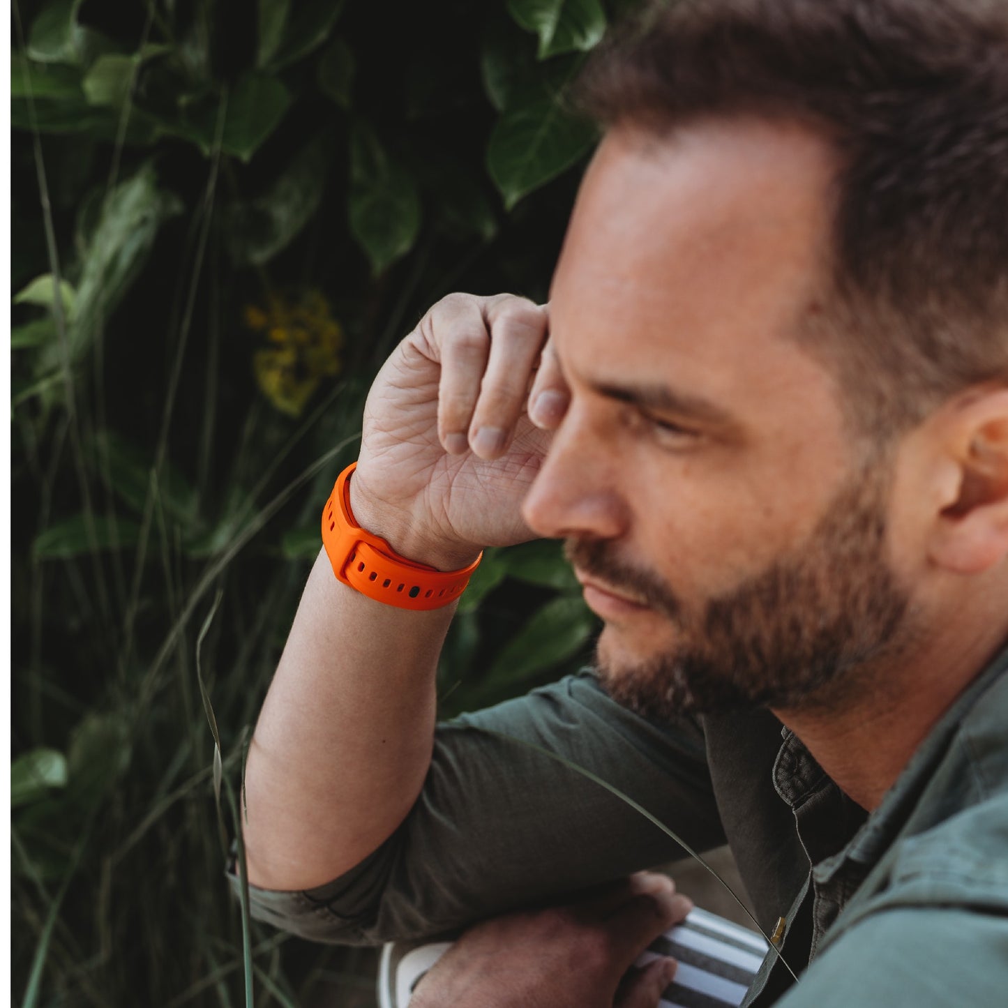 Ein Mann mit kurzen braunen Haaren und Bart trägt ein arktisband Apple Watch FKM Sportarmband "Sweat" von arktisband, dazu ein orangefarbenes Armband und ein grünes Hemd. Er stützt sein Kinn auf seine Hand. Im Hintergrund ist üppiges grünes Laub zu sehen.