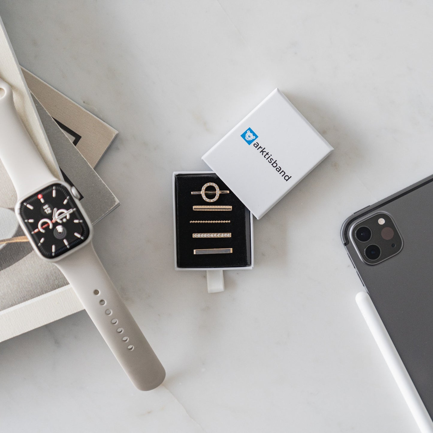Ein arktisband Apple Watch Charm „O Style“ in Beige, ein vergoldetes Ringset in einer Box und ein Smartphone auf weißem Marmor – modische Accessoires von arktisband für jeden Anlass.