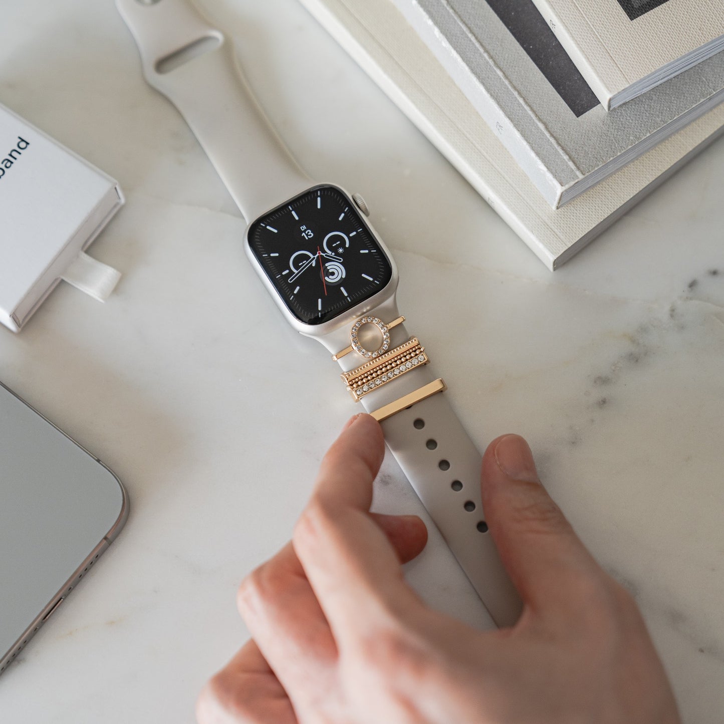Eine Hand berührt eine Apple Watch mit einem beigen Arktisband und Arktisband Apple Watch Charms „O Style“ auf einer Marmoroberfläche.
