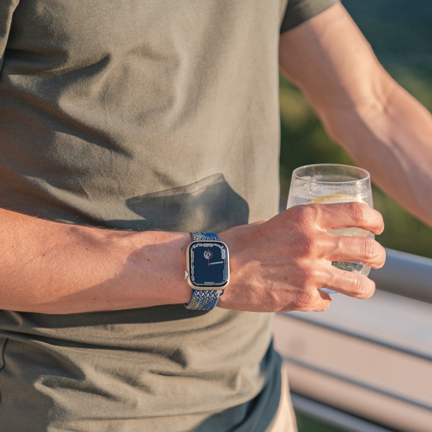 Eine Person trägt ein geflochtenes Flex Loop TwoTone Armband von Arktisband für die Apple Watch und hält ein klares Glas Wasser mit einer Zitronenscheibe in der Hand. Das blaue Armband von Arktisband passt zu ihrem kurzärmeligen olivgrünen Hemd.