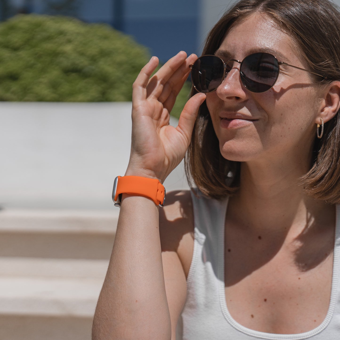 Eine Frau mit Sonnenbrille und weißem Tanktop sitzt lächelnd im Freien und präsentiert das arktisband Apple Watch Silikonarmband „Aqua“ von arktisband an ihrem linken Handgelenk. Im Hintergrund sind Grünflächen und Treppenstufen zu sehen.
