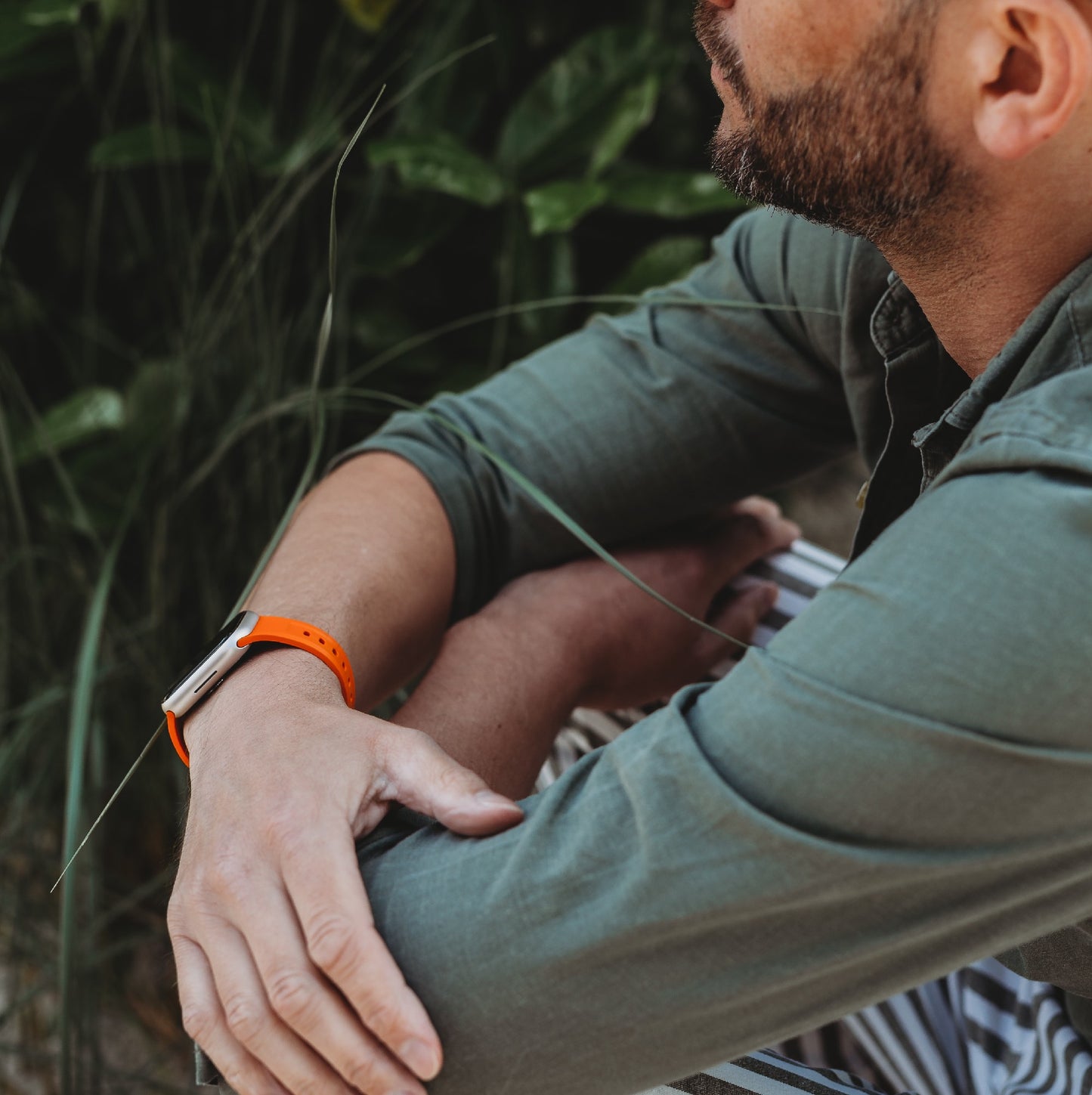 Eine Person mit gestutztem Bart sitzt im Freien, trägt ein grünes Hemd, trägt das arktisband Apple Watch FKM Sportarmband „Sweat“ von arktisband und ruht mit den Armen auf den Knien inmitten von grünem Laub.