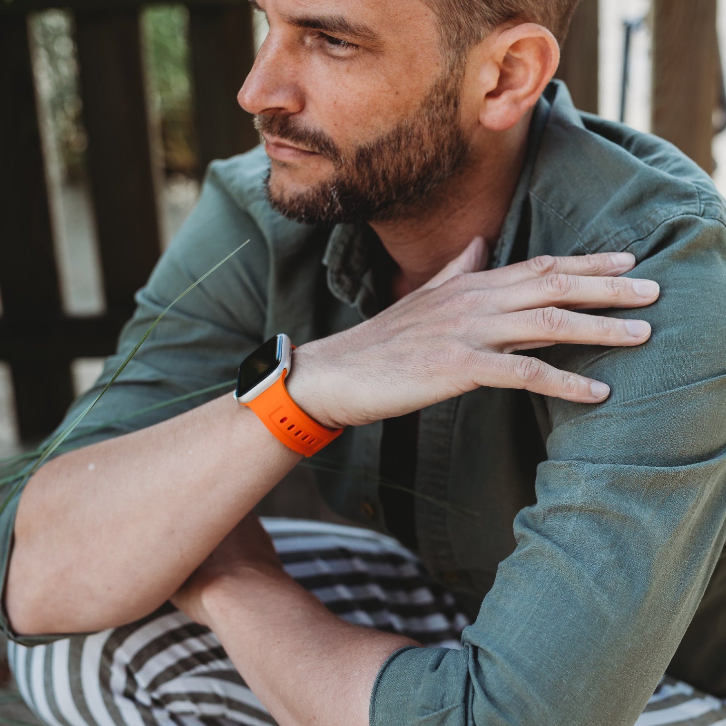 Ein Mann mit gestutztem Bart und kurzen Haaren in einem grünen Hemd und gestreiften Hosen sitzt mit verschränkten Armen da, trägt das orangefarbene arktisband Apple Watch FKM Sportarmband „Sweat“ von arktisband und blickt in die Ferne.