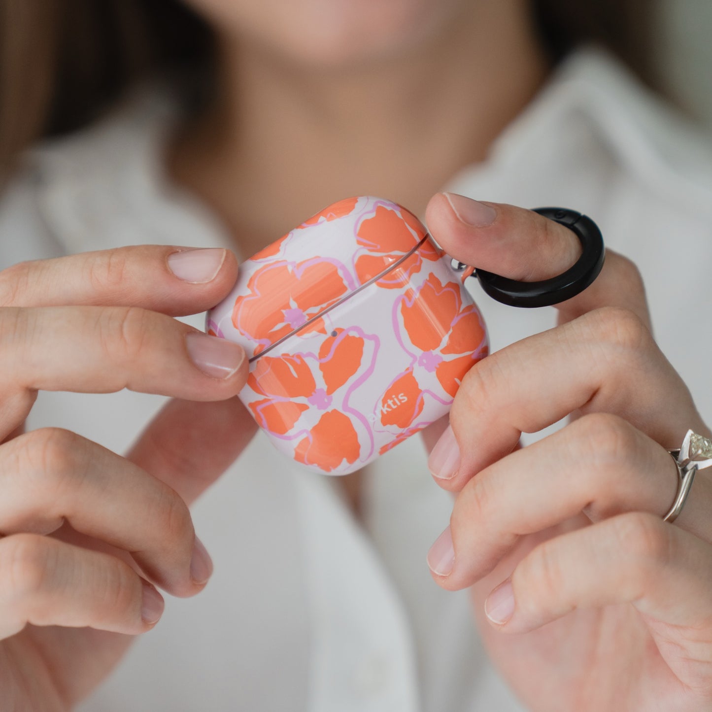 Hände, die eine Arktis AirPods 5-Hülle – Orange Mallow – mit Blumenmuster und einem daran befestigten schwarzen Karabinerhalter halten.