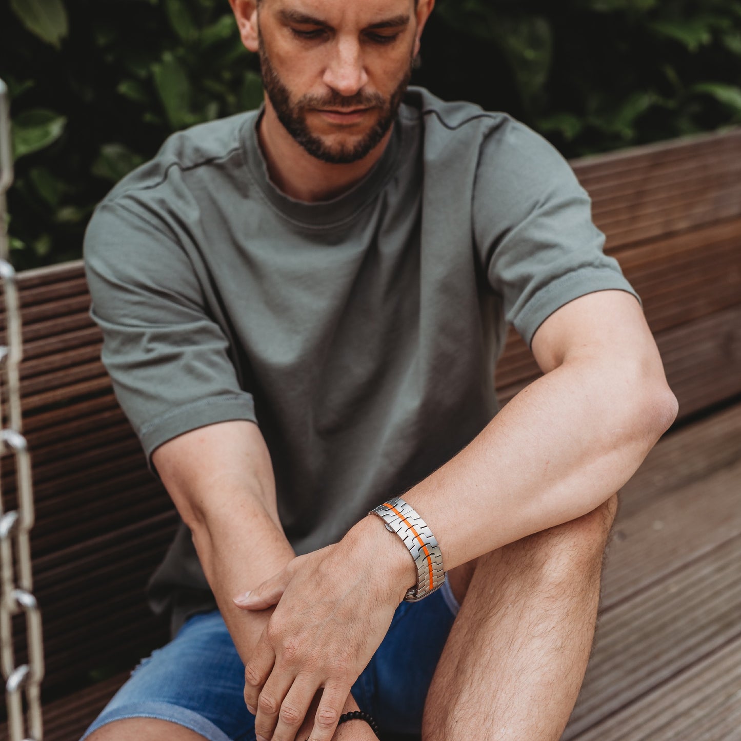 Ein Mann in grauem T-Shirt und blauen Shorts sitzt draußen auf einer Holzbank und blickt nach unten. Sein Arm ruht auf seinem Knie. Er trägt das Arktisband Apple Watch Titan Armband „Aviato Edition“. Im Hintergrund ist grünes Laub zu sehen.